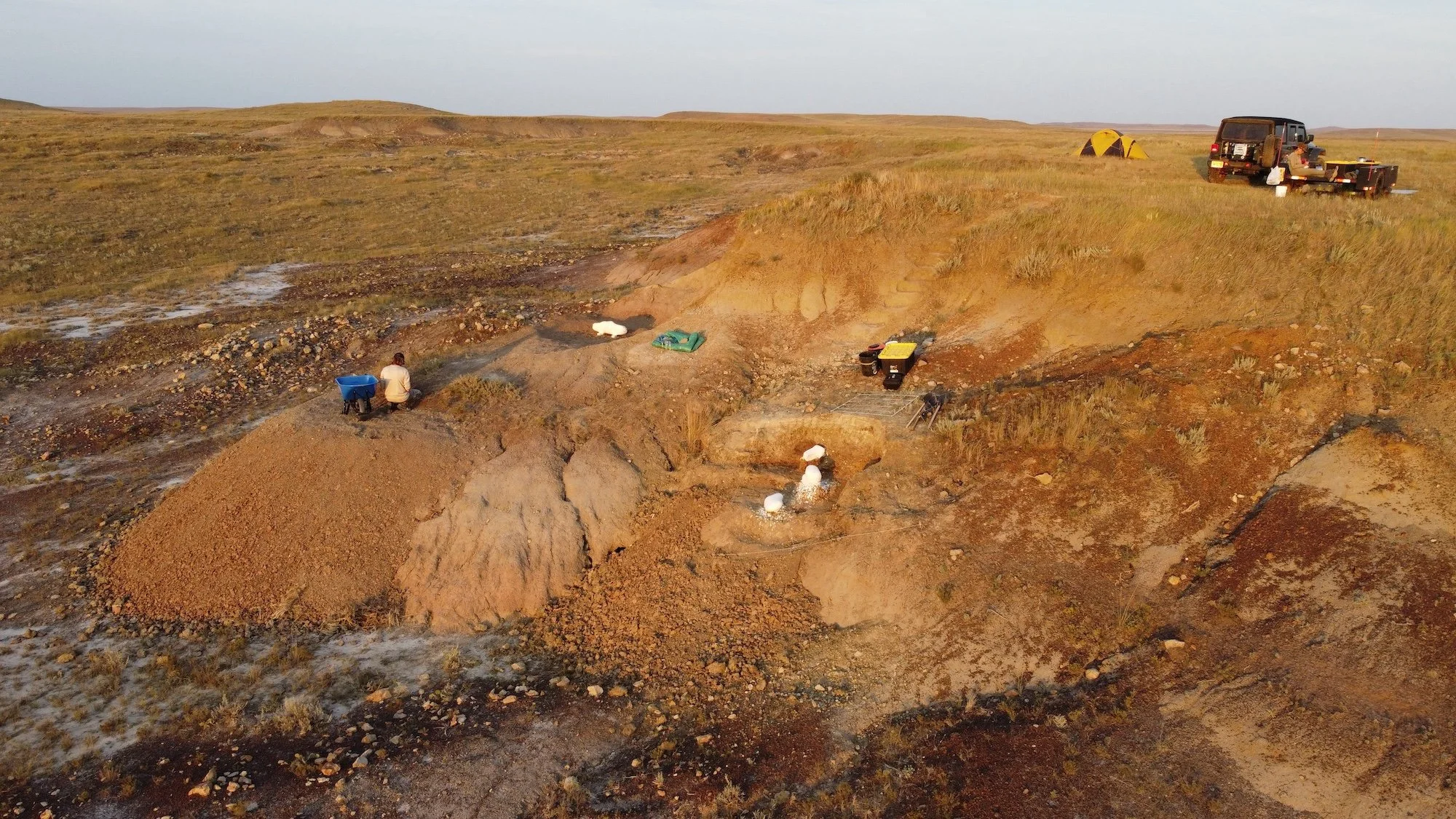 A person is sitting on a small hill of dirt in a vast, open landscape with grass, rocks, and small patches of water. Nearby, there are containers, equipment, and a vehicle with a trailer. In the background, a yellow tent and a large black vehicle are
