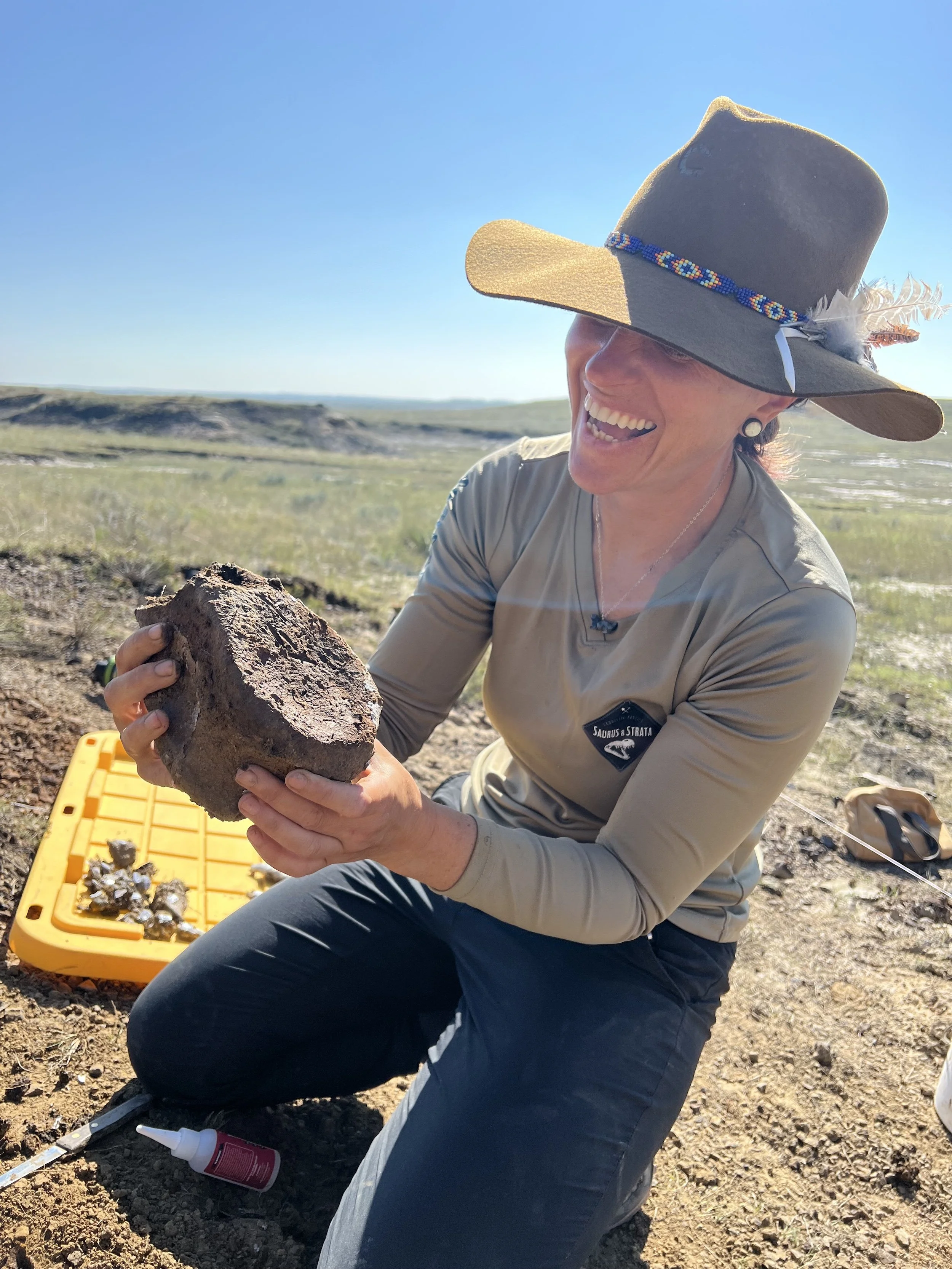 A woman wearing a wide-brimmed hat, earrings, and outdoor clothing is smiling and holding a large rock or fossil in an open field with a clear blue sky.