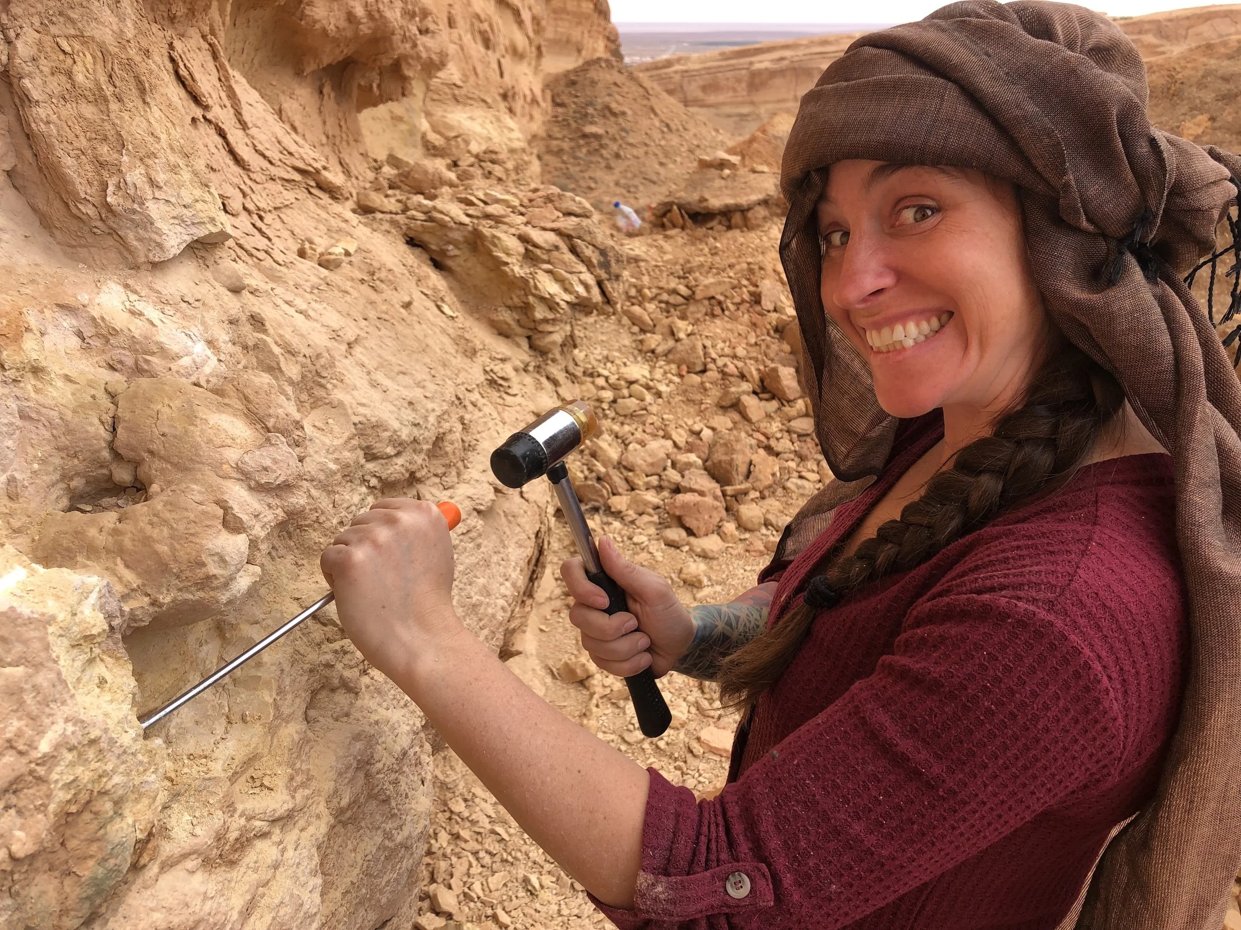 A woman with a braid, wearing a brown head covering and a maroon sweater, smiling while examining a rock face with a hammer and chisel in a desert landscape.