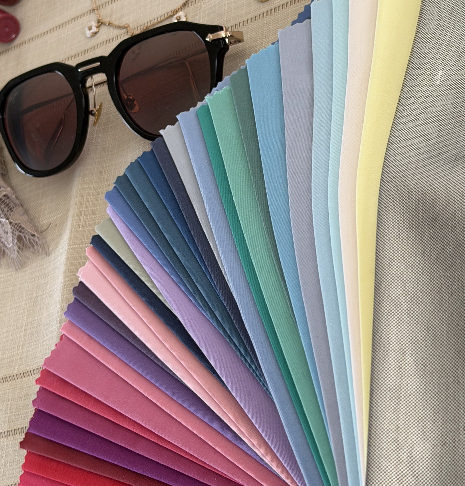 A pair of black sunglasses with round lenses resting on a pile of fabric swatches in various pastel and vibrant colors, arranged in a fan shape on a beige tablecloth.