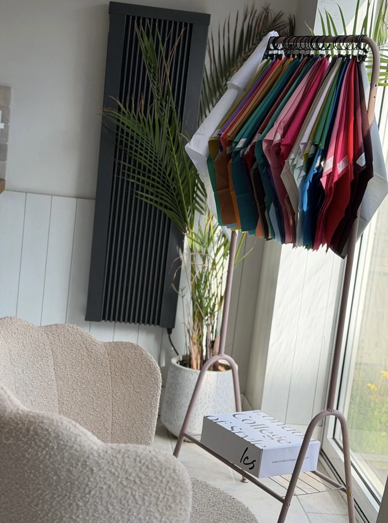 Indoor setting with a clothing rack holding colorful fabric swatches, a tall black radiator, a potted plant, a beige textured chair, and a box on a small pink table near a large window.
