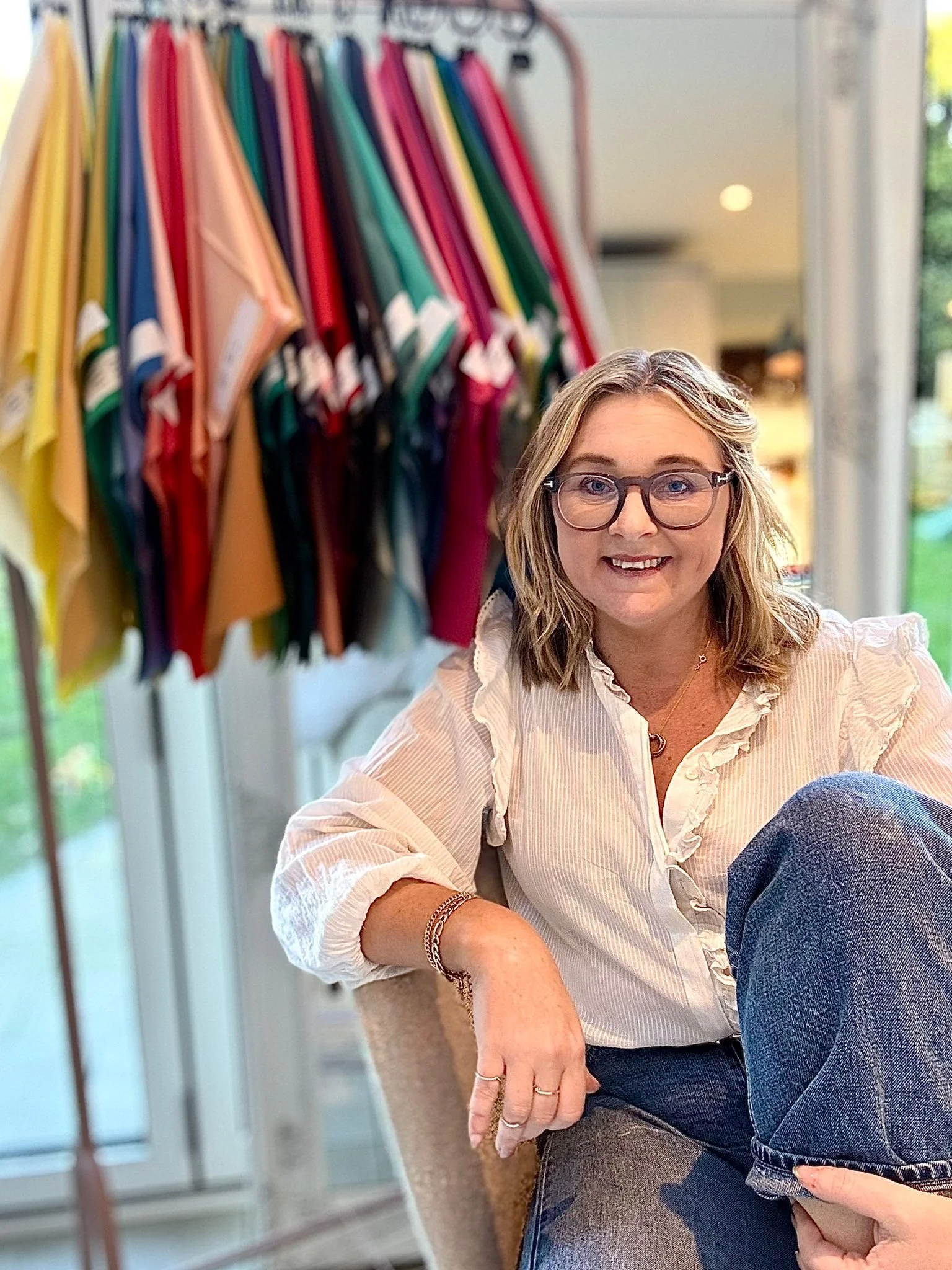 A woman with glasses and blonde hair sitting indoors, smiling at the camera, with colorful fabric swatches on a rack behind her.