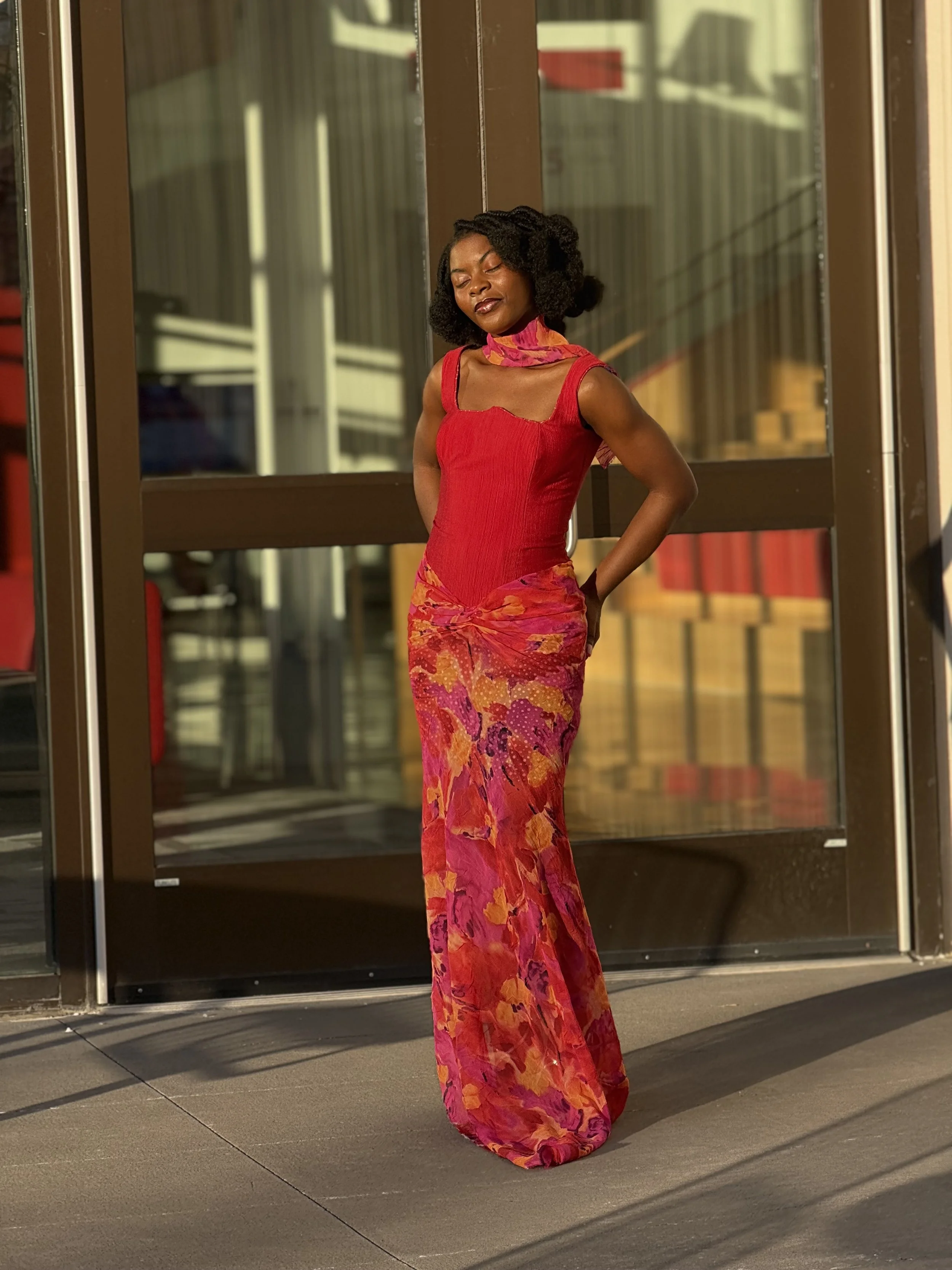 A woman in a red top and a long, colorful floral skirt standing outdoors in front of glass doors with her eyes closed and hands behind her back.