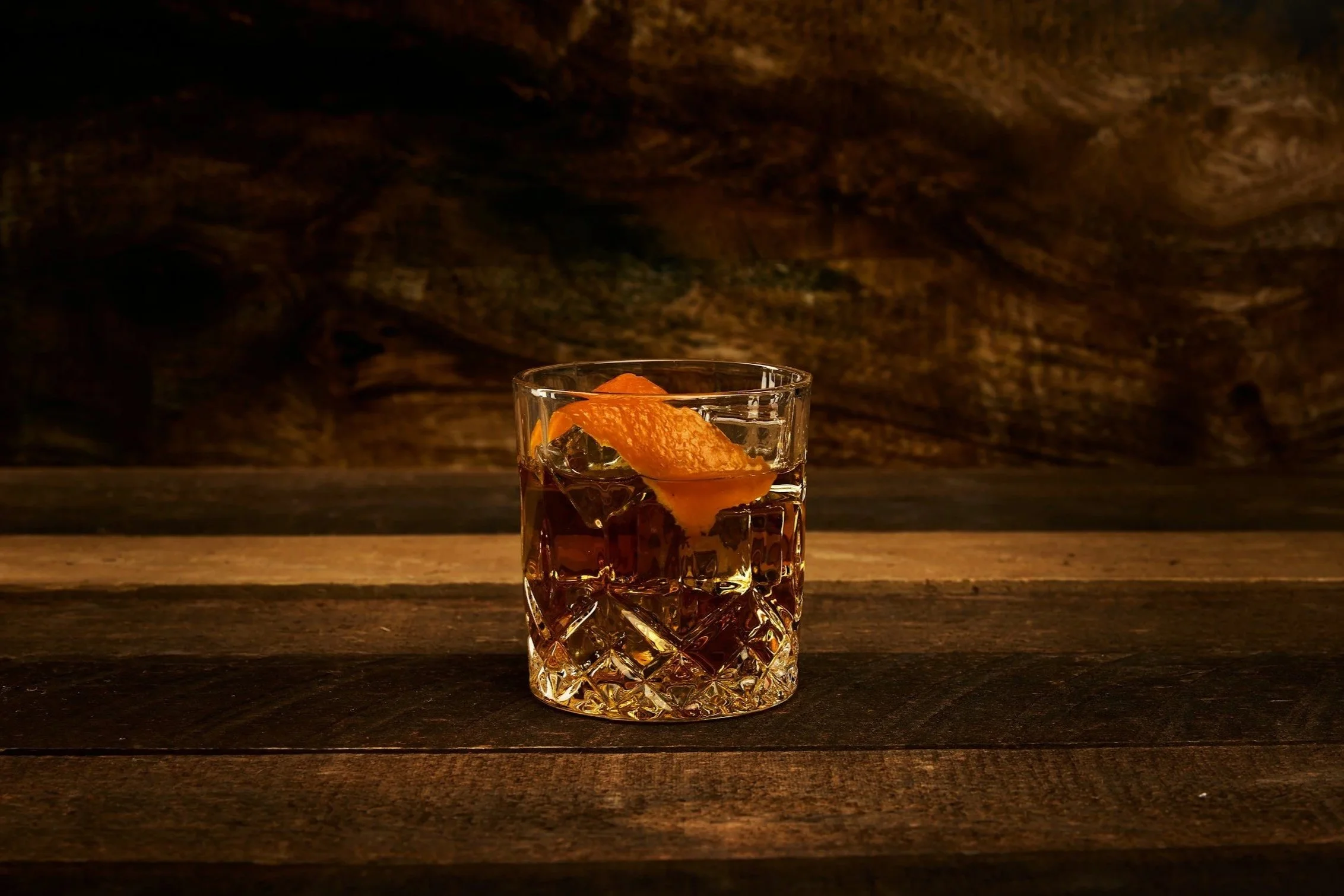 A glass of dark liquor with ice and an orange peel garnish on a wooden surface, dark background.