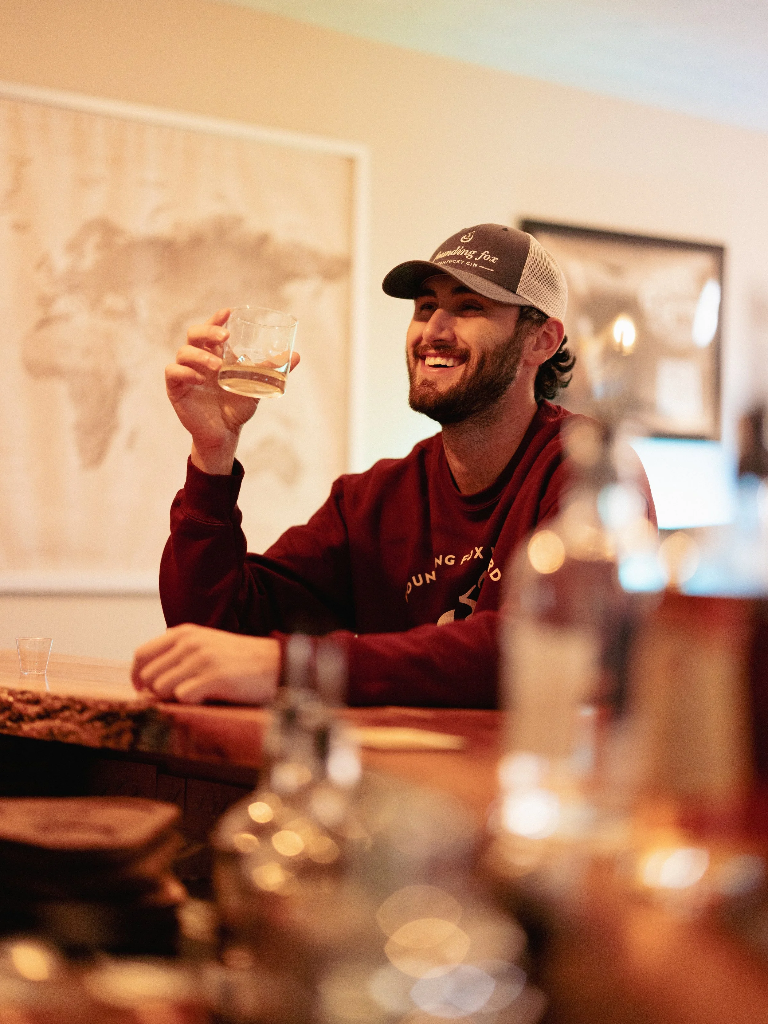 A young man with a beard, wearing a cap and a maroon sweatshirt, is sitting at a bar holding a glass of whiskey, smiling, with a world map on the wall behind him and bottles on the counter in the foreground.