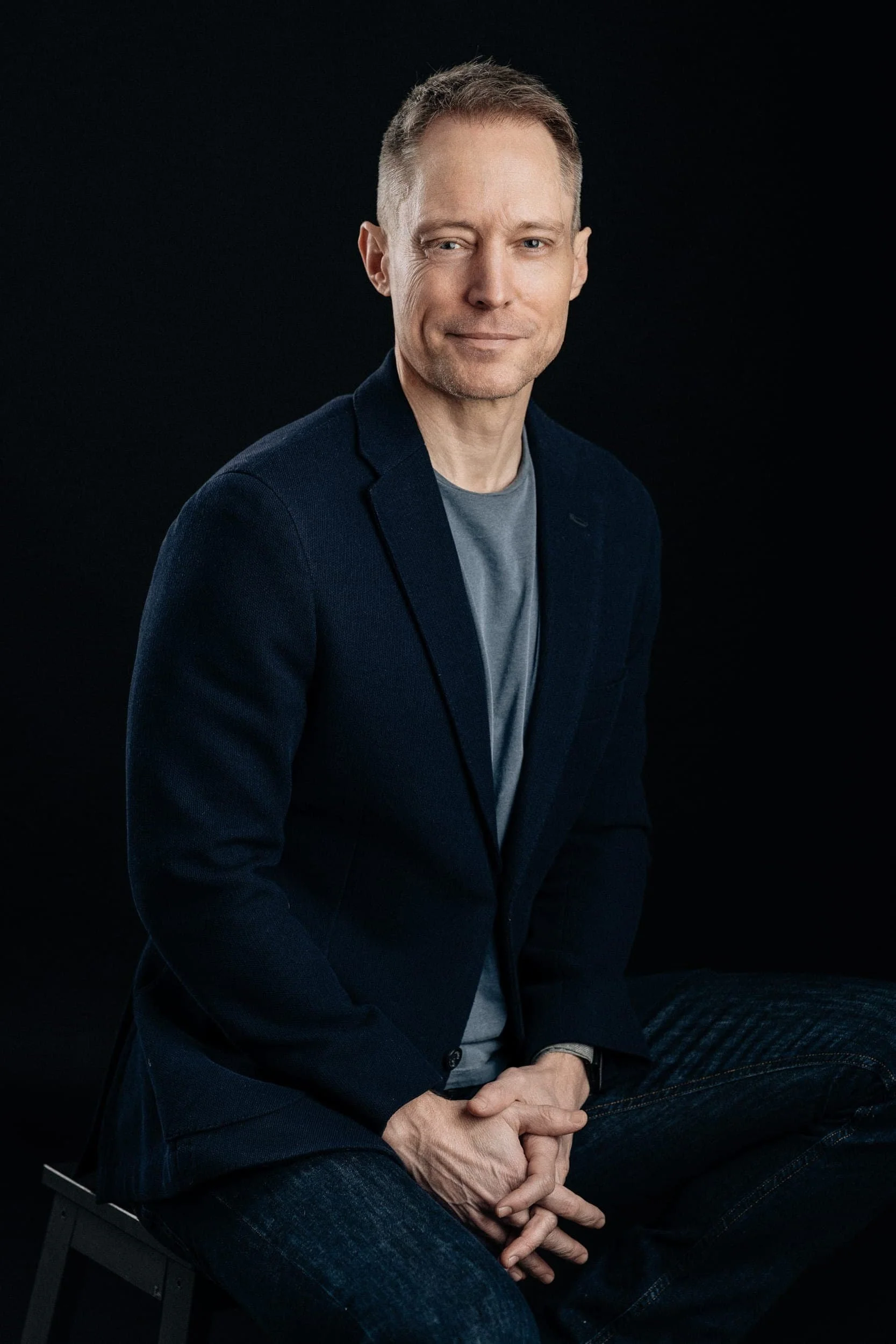 A man in a navy blazer and blue jeans sits on a stool against a dark background, smiling with hands clasped in his lap.
