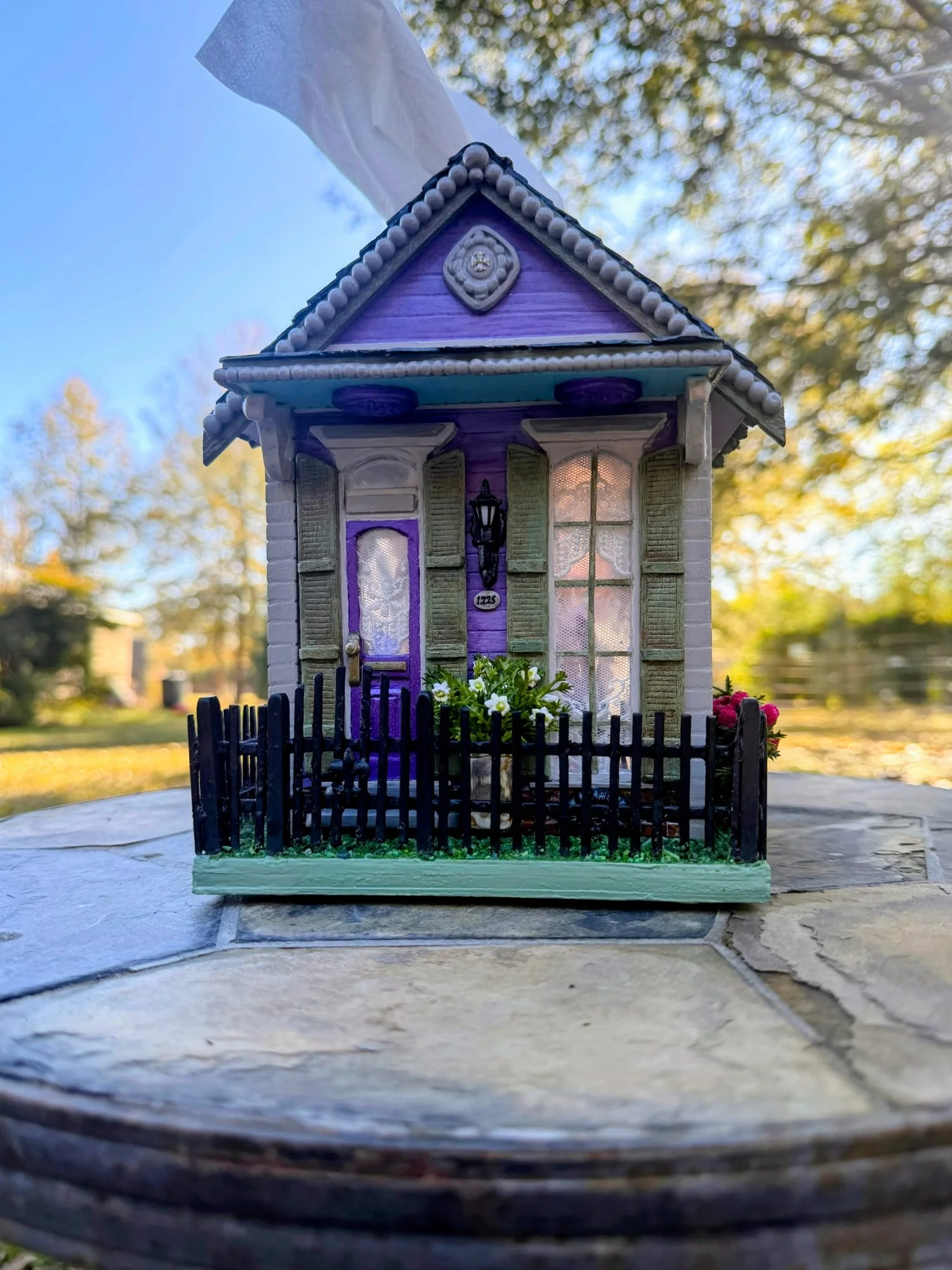 Front view of the Purple Reign Shotgun House diorama by Art Illusions by Young. Lavender facade with gable roof, porch overhang, decorative slate roof tiles, and golden cross on the sculpted facade carving. The black wrought iron fence and blooming y