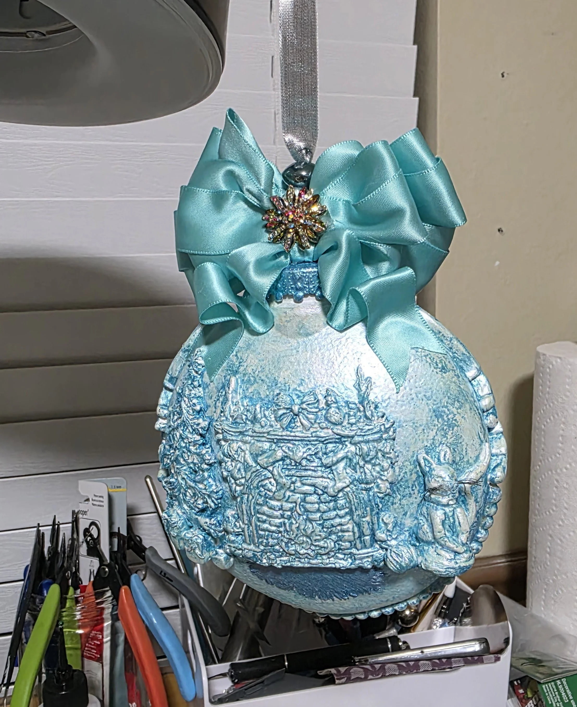 Studio view of the Cozy Fireplace with Cat Ornament by Art Illusions by Young. The clay enhanced spherical form displays its intricate and waxed light blue texture, crowned by a large aqua satin ribbon bow. Generously sized and luminous, this ornamen