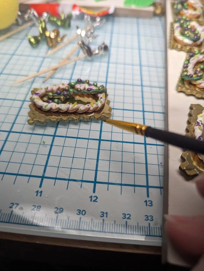 Studio process shot of a Mardi Gras King Cake Baby magnet/pin by Art Illusions by Young. An artist’s brush applies details to the clay tray, bringing authentic detail and color to the miniature sculpture. before it is framed with gold foil and Carniv