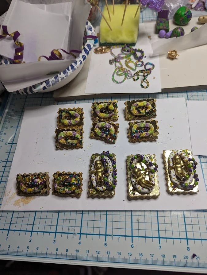 Studio display of Mardi Gras King Cake Baby magnets/pins by Art Illusions by Young. A collection of both hand-sculpted and painted babies in sculpted king cakes with colorful bead accents in traditional Carnival colors sits alongside additional workb