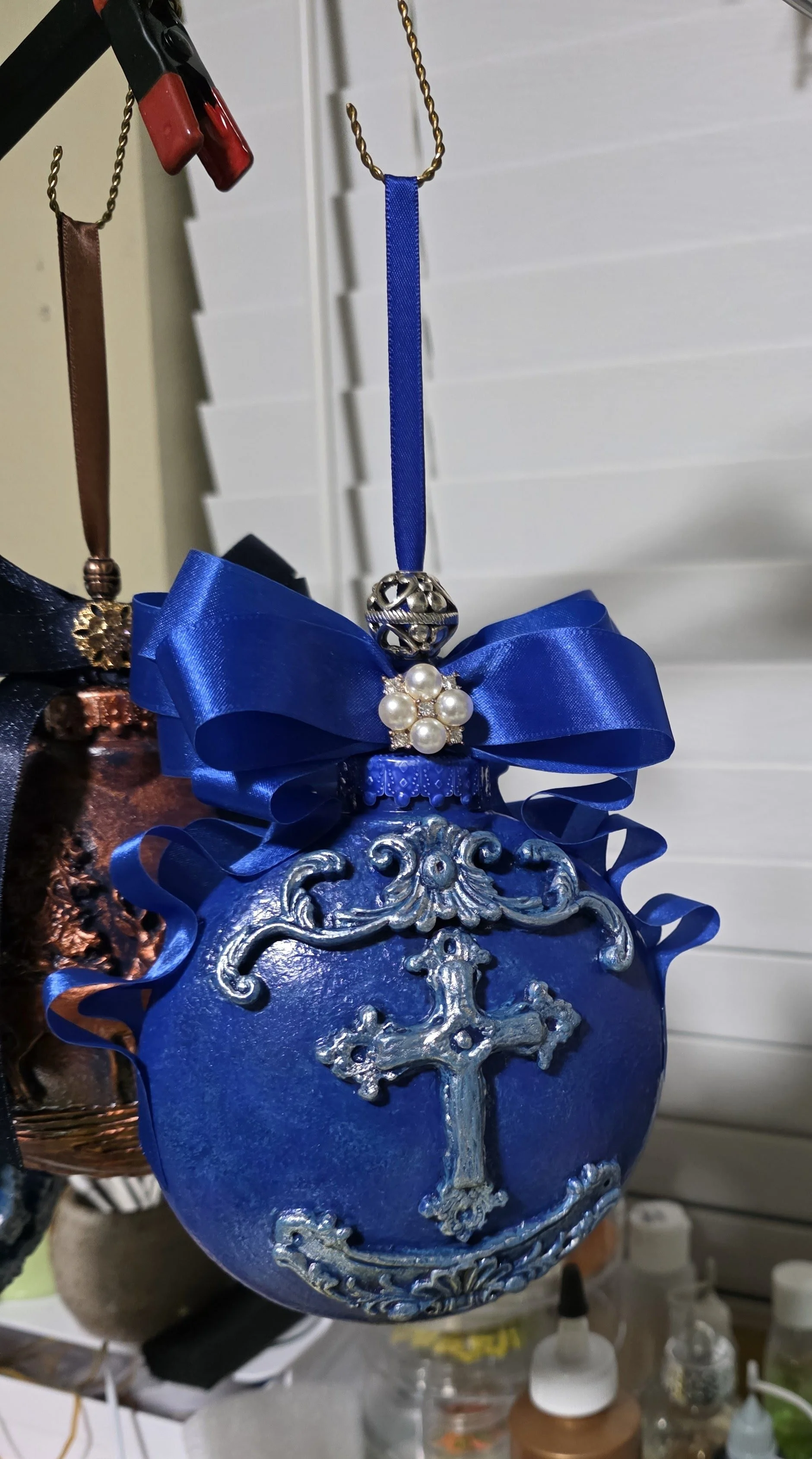 Studio view of the Royal Blue Cross Ornaments by Art Illusions by Young before adding the European faceted crystal embellishments. The ornament is suspended over the workbench, revealing the luminous hand waxed finish, ornate silver cross and scrollw