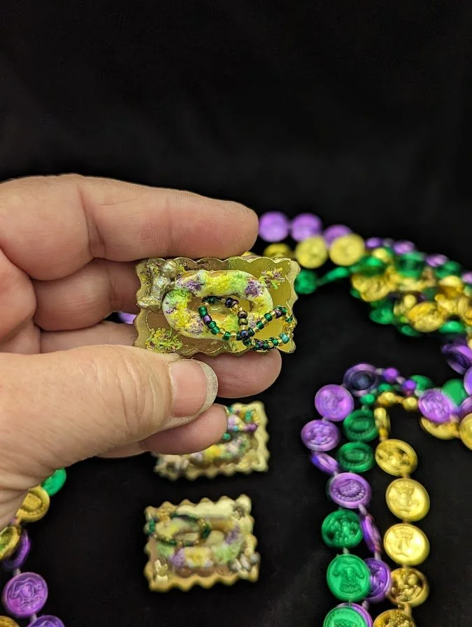 Hand holding a Mardi Gras King Cake Baby pin by Art Illusions by Young. The miniature hand-sculpted baby, nestled in a tiny hand-sculpted cake with a purple, green, and gold seed beads necklace, showcases the charming sculpted details and authentic C