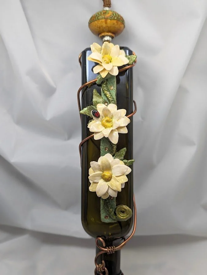 Handcrafted decorative hummingbird feeder by Art Illusions by Young with a green glass body, adorned with polymer clay white flowers, green leaves, a small ladybug, copper wire accents, and a spherical ceramic and green finial.