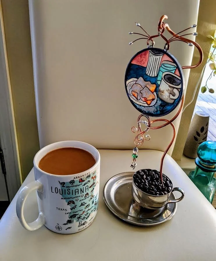 Café au Lait Illusion Glass displayed on its custom handcrafted stand.
This warm Louisiana-themed piece features a hand-painted Cafe' au Lait Illusion Glass suspended from swirling copper “steam” above a vintage silver-plated cup and saucer filled wi