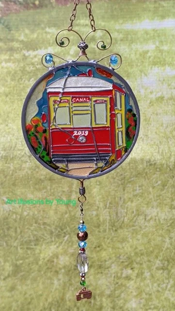 🔴 Sold: 
Red New Orleans Streetcar Illusion Glass by Art Illusions by Young. The iconic red streetcar glides along its tracks in this vibrant circular suncatcher. Hand-painted architectural details and warm lighting evoke the romance and energy of N