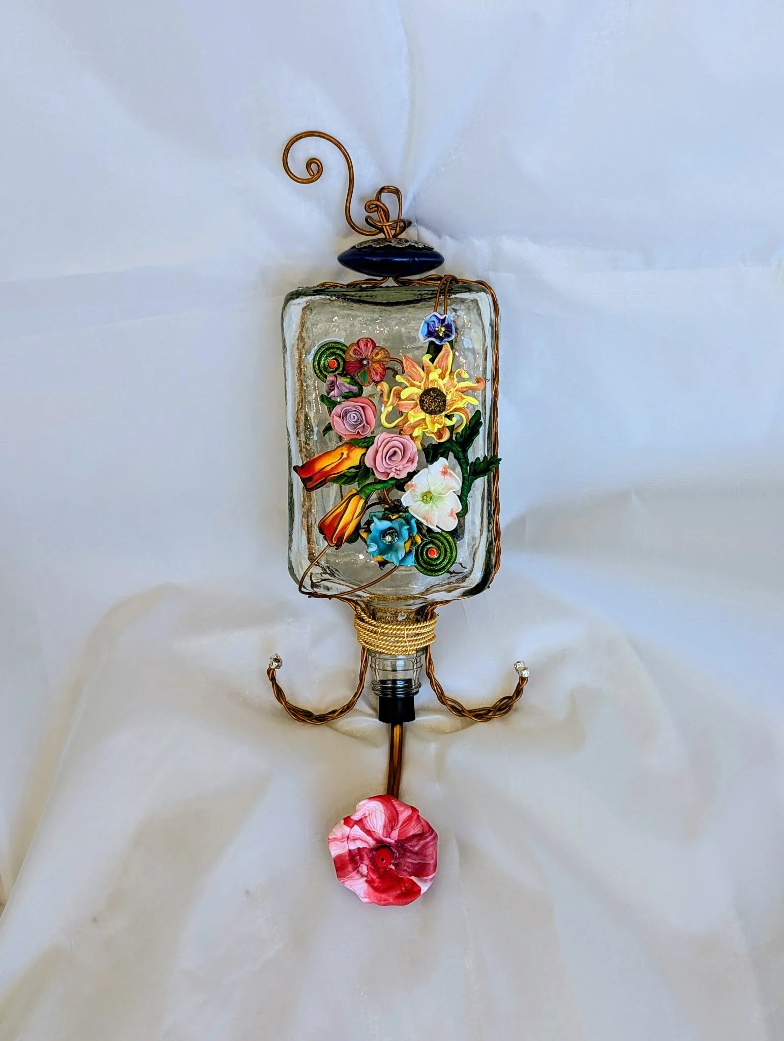🔴 Sold: 
Lantern-Style Floral Hummingbird Feeder with Red and Yellow Accent by Art Illusions by Young, Folsom, Louisiana. This charming lantern-shaped glass feeder is adorned with a profusion of hand-sculpted polymer clay flowers in bright, multicol