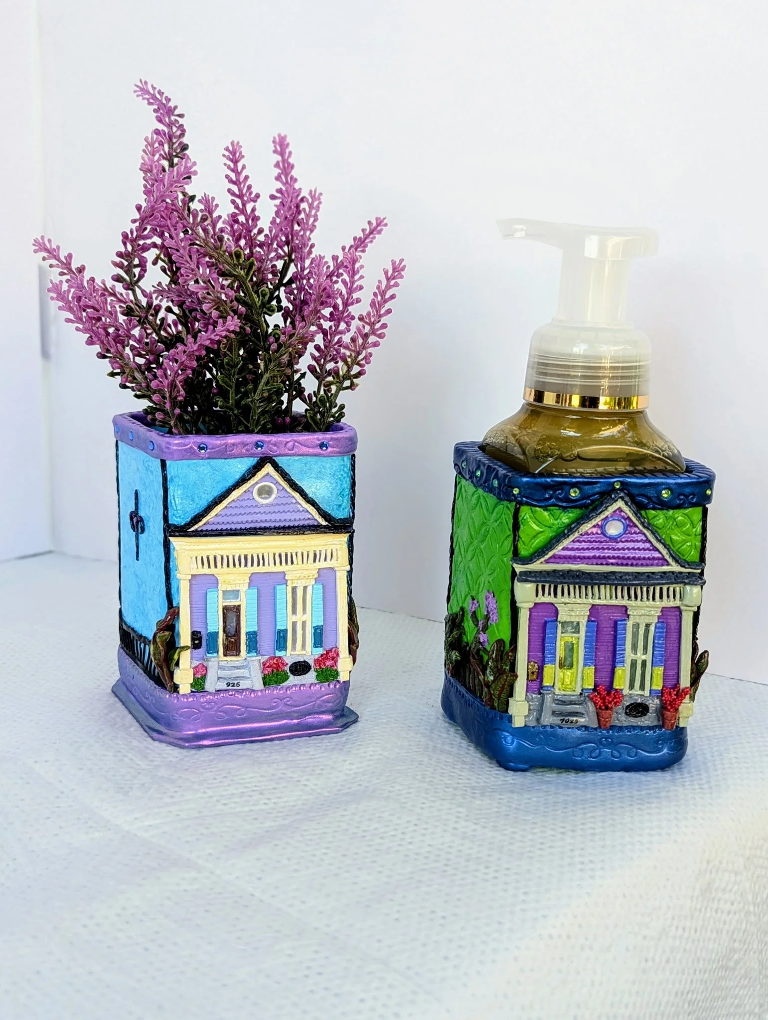 These handcrafted functional art pieces by Art Illusions by Young capture the vibrant charm of historic Creole shotgun houses. One features a soft lavender facade with sage-green shutters and ornate gingerbread trim; the other displays a rich green e
