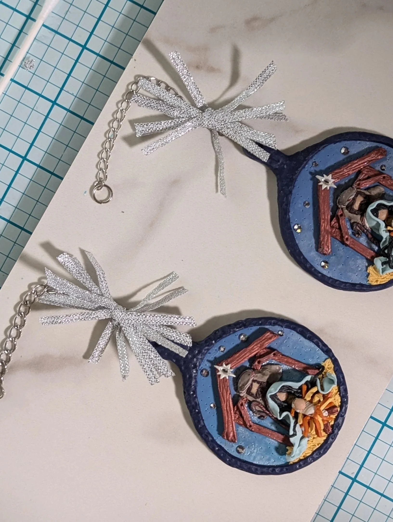 Studio view of two Nativity Manger Ornaments by Art Illusions by Young shown with silver starburst ribbon bows and a ruler for scale. The hand-sculpted manger scenes — complete with Mary, Joseph, the infant Jesus in golden hay, and a simple stable ro