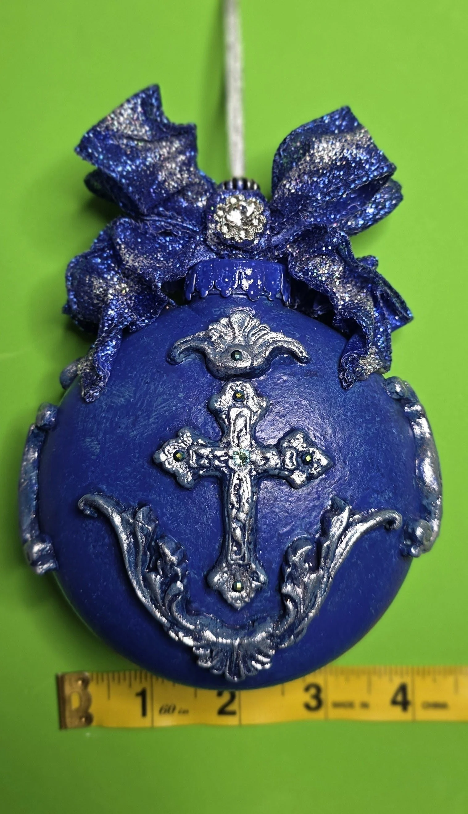 Measured view of the Celestial Blue Cross Ornament by Art Illusions by Young. The luminous cobalt blue surface and intricate silver cross with raised flourishes are visible from multiple angles, while the sparkling royal-blue and silver ribbon bow ad