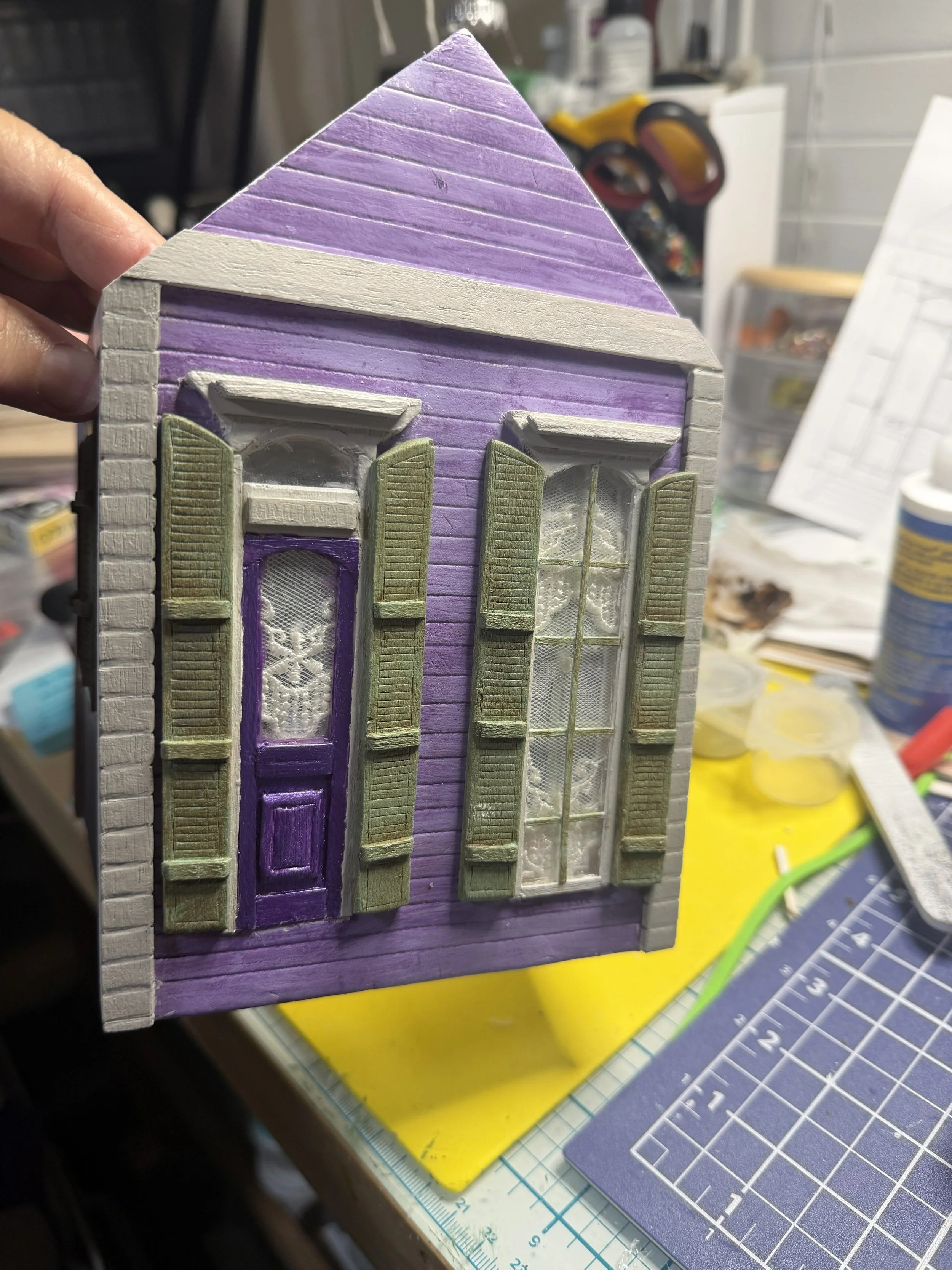 Front wall detail of the Purple Reign Shotgun House diorama by Art Illusions by Young. Sage green shutters frame reflective acetate windows with lace curtains, while wood-burned details are featured on the aged sage shutters. Architectural elements a