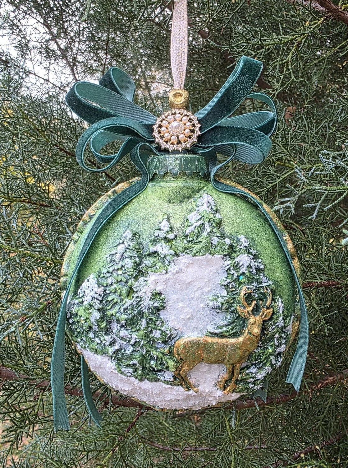 Frontal close-up of the Woodland Majesty Ornament by Art Illusions by Young on a green background. The majestic stag stands amid snow-dusted trees and foliage in shimmering metallic green tones, highlighted by a generous dark green velvet ribbon bow 