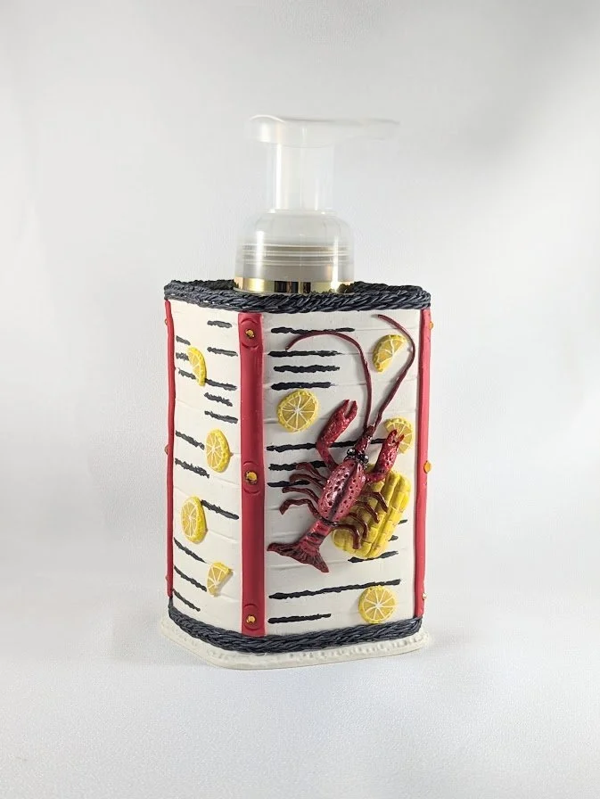 🔴 Sold: 
Crawfish Soap Dispenser by Art Illusions by Young. A fun and functional soap dispenser covered in hand-sculpted polymer clay depicting a lively Cajun crawfish boil with corn, lemons, and newspaper texture. The vibrant red and yellow details