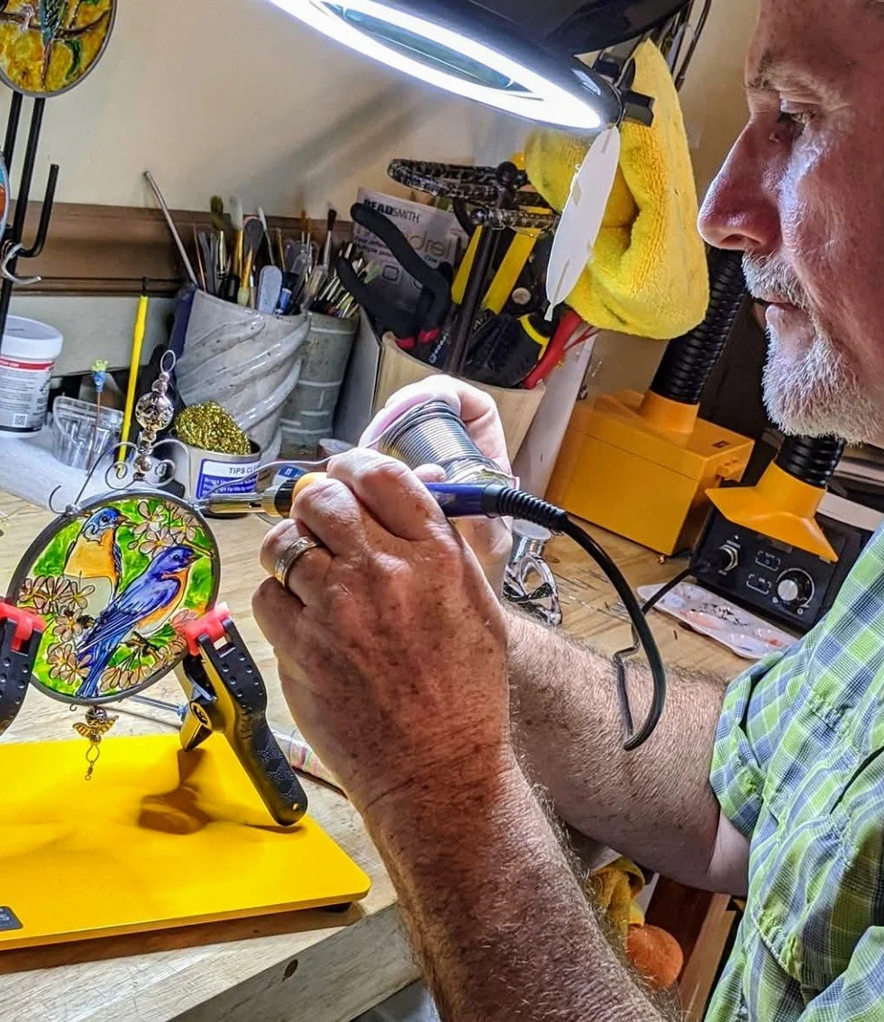 Mark Young using a soldering iron to assemble a stained glass ornament with blue birds and flowers, part of the handcrafted Illusion Glass process at Art Illusions by Young