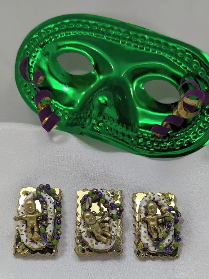 Green Mardi Gras mask surrounded by king cake baby pins by Art Illusions by Young. The  painted babies, each on ornate gold-foiled trays with hand-sculpted clay cake and purple, green, and gold bead accents, complement the festive mask. These versati