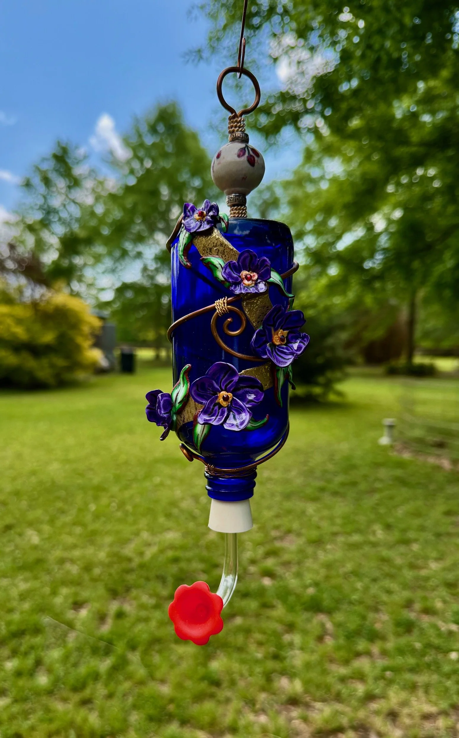 Handcrafted hummingbird feeder by Art Illusions by Young featuring colorful glass and decorative elements, hanging outdoors with trees and sky visible in the background.

