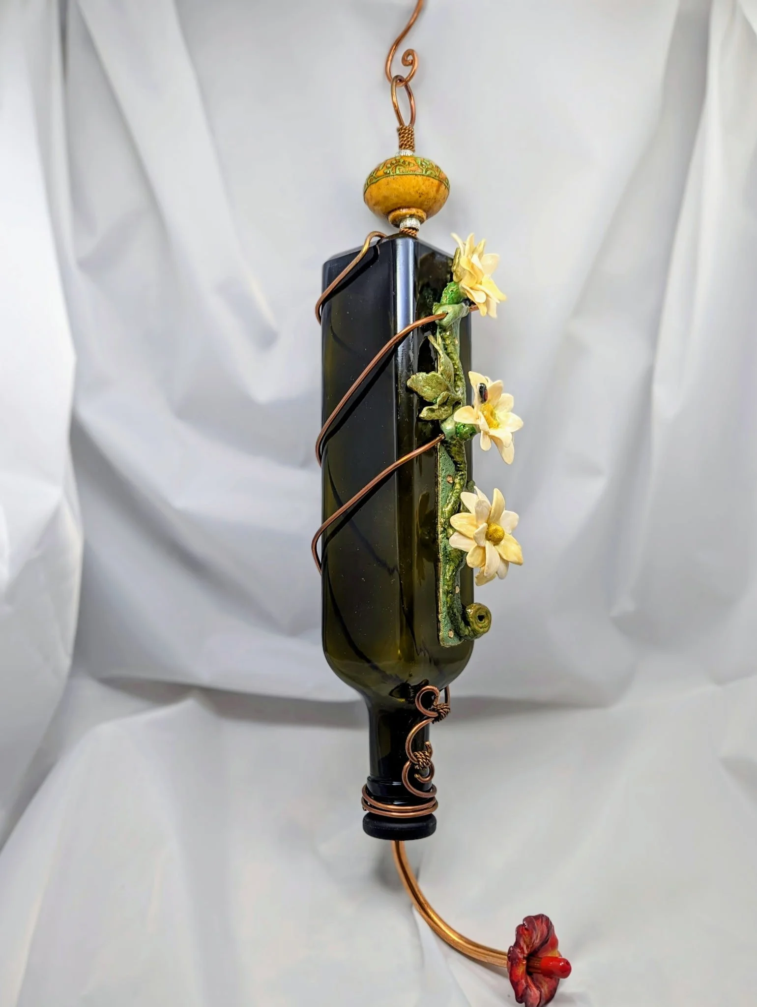 Handcrafted decorative hummingbird feeder by Art Illusions by Young featuring a green rectangular bottle entwined with copper wire, adorned with cream-colored flowers, green leaves, a yellow and green ceramic knob, and copper hanger.