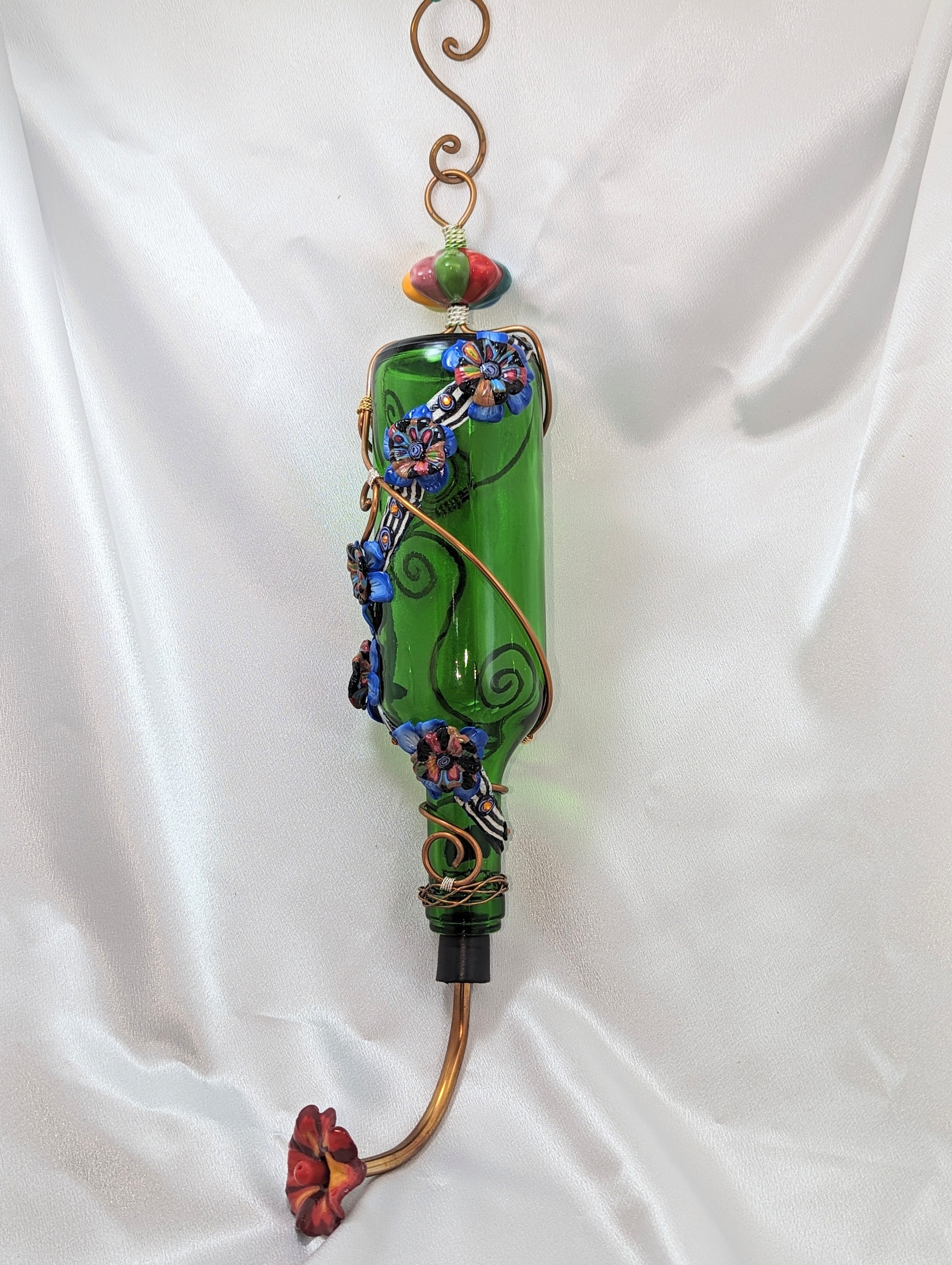 Right side view of the Emerald Garden Bloom Hummingbird Feeder by Art Illusions by Young. The emerald green glass bottle is wrapped in decorative copper wire vines, accented with meticulously hand-sculpted polymer clay flowers in rich blue and multic