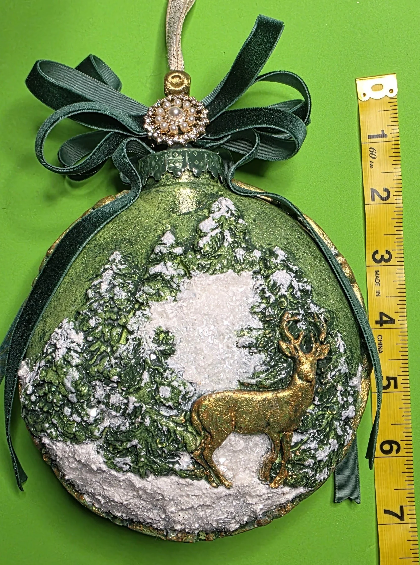 Measured view of the Woodland Majesty Ornament by Art Illusions by Young with ruler for scale. The hand-sculpted spherical ornament displays the stag and winter forest scene in rich metallic green and gold, crowned by a dark green ribbon bow. Compact