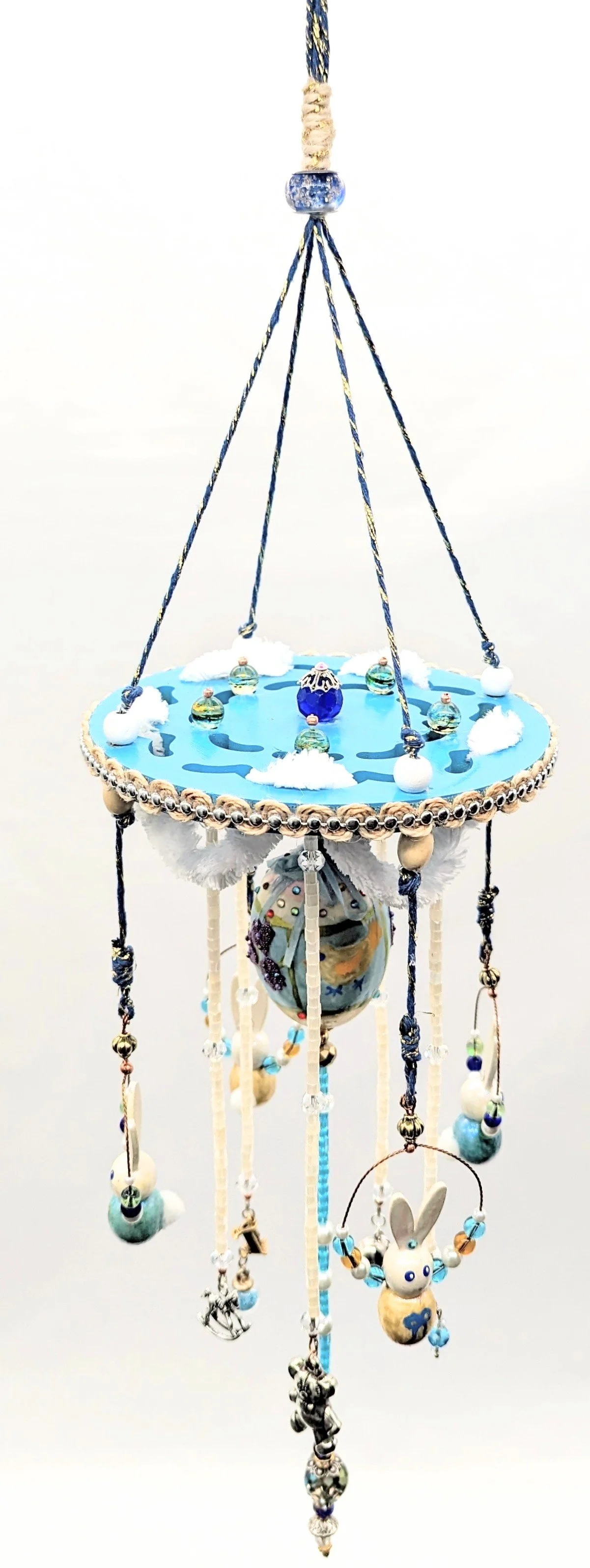 Top view of the Baby Mobile – Cherished Nursery Keepsake by Art Illusions by Young. The painted blue wooden disc with scalloped white fluffy cording and golden accents forms the centerpiece, from which hand-painted wooden bunnies and the decorated bl