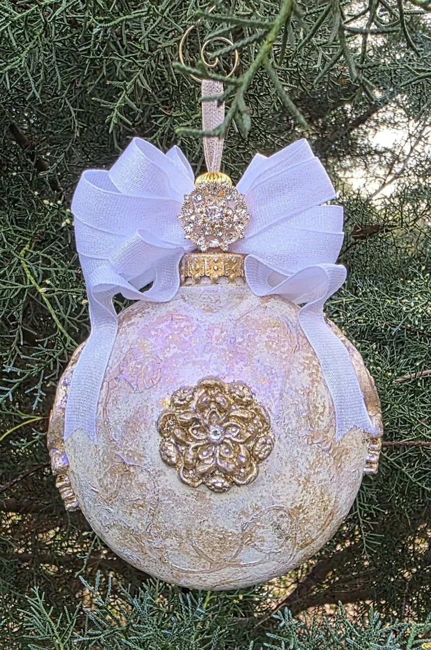 A close-up hand-sculpted and molded Guardian Angel Ornament by Art Illusions by Young, viewed from the back while hanging among evergreen branches. The smooth, luminous iridescent pearl and blush surface displays subtle golden accents and delicate te