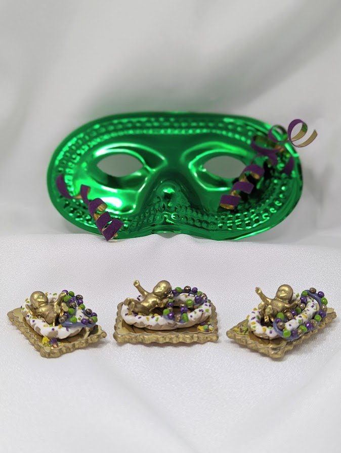 Green Mardi Gras mask paired with three king cake baby pins by Art Illusions by Young. The hand painted babies feature intricate and gold-foiled hand-sculpted cakes and colorful beadwork in purple, green, and gold. These lightweight, versatile pieces