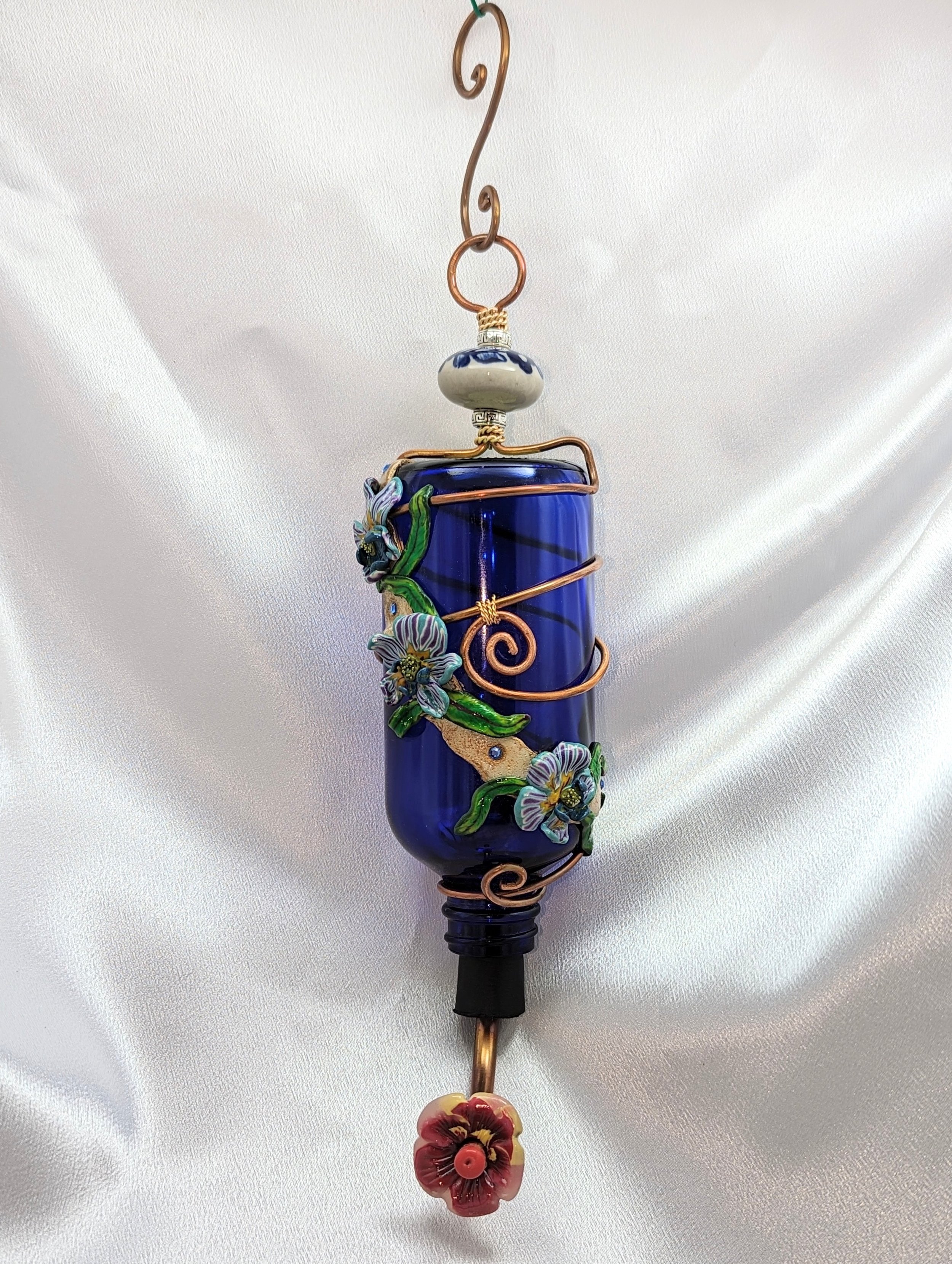 🔴 Sold: 
Cobalt Blue Hummingbird Feeder with Greenery by Art Illusions by Young, Folsom, Louisiana. A striking cobalt blue glass bottle adorned with hand-sculpted polymer clay vines and leaves in lush greens, accented by intricate copper wire wrappi