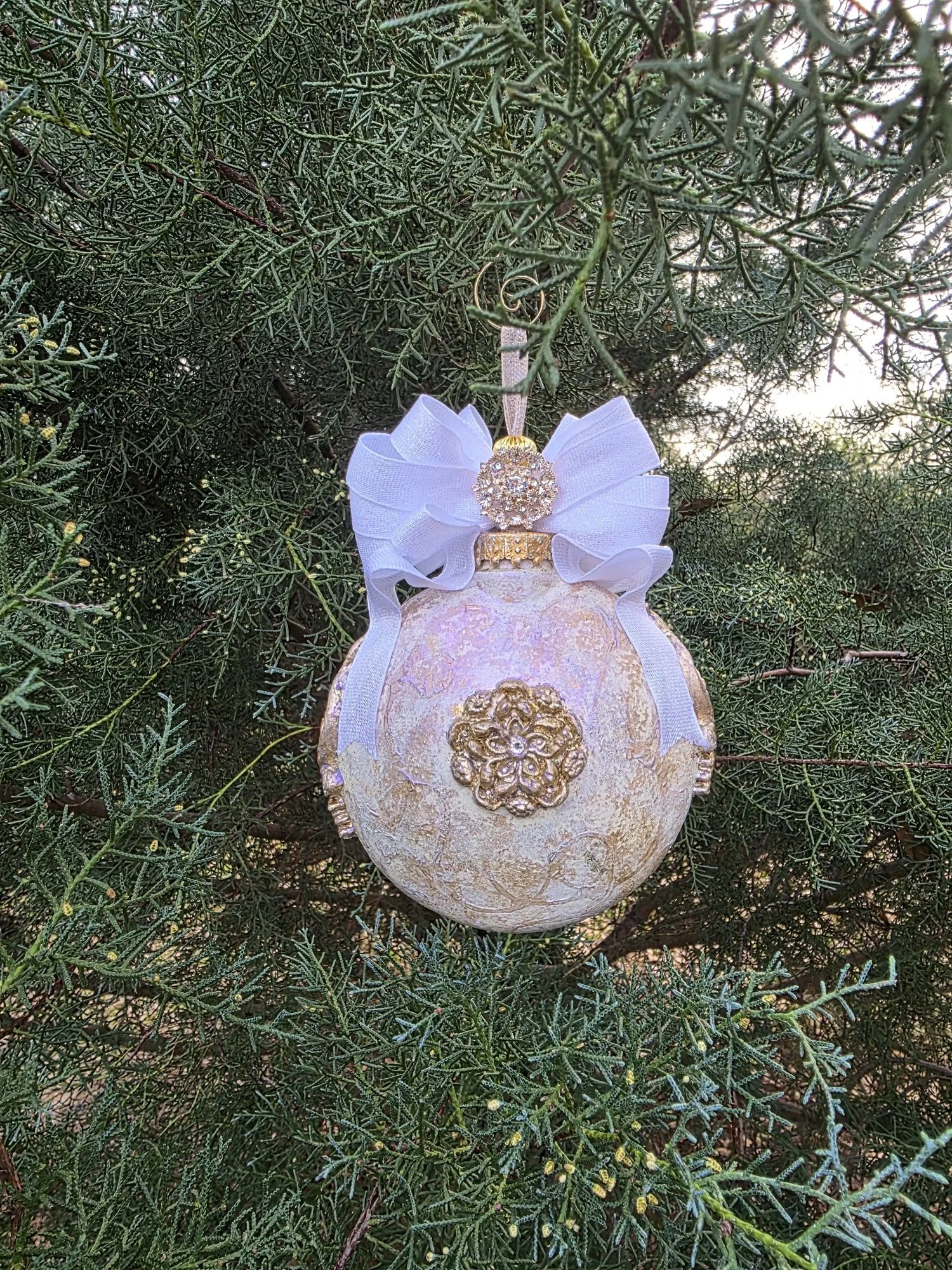 A hand-sculpted and molded Guardian Angel Ornament by Art Illusions by Young, shown from the back while hanging among lush evergreen branches. The reverse side reveals the smooth, iridescent pearl and blush finish with subtle golden accents and delic