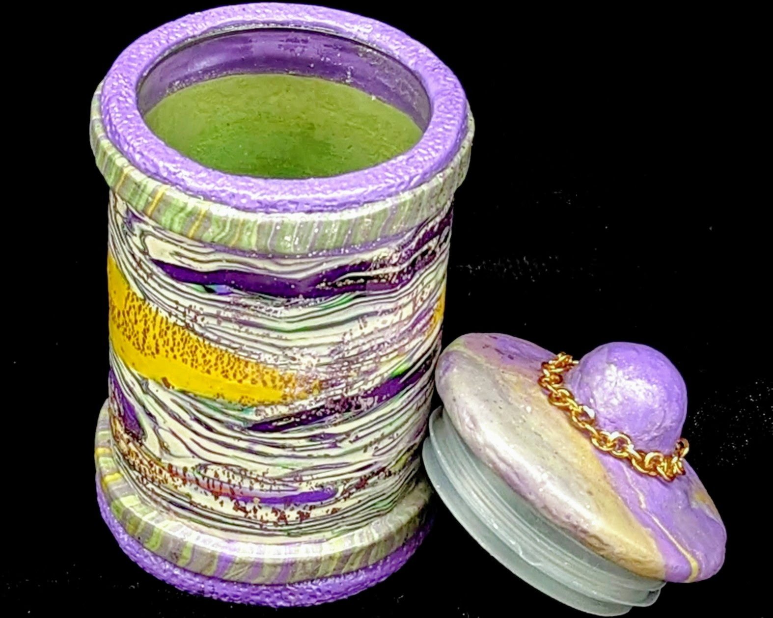 🟢 Available :
Carnival Opulence Lidded Vessel by Art Illusions by Young, Folsom, Louisiana. This sculptural polymer clay jar displays intricate marbled layers of amethyst purple, emerald green, gold, and soft white with a soft purple and gold marble