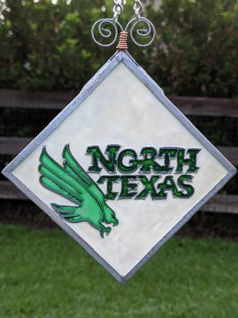 🔴 Sold: 
North Texas Illusion Glass by Art Illusions by Young. A bold diamond-shaped suncatcher proudly displays the North Texas emblem with a green eagle and clear lettering. The strong design and vibrant colors make this a striking tribute to regi