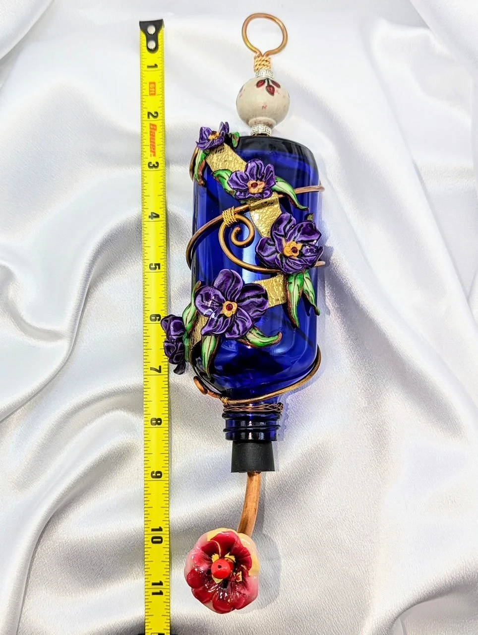 Measured view of the Hummingbird Feeder
Purple Pansy Elegance by Art Illusions by Young with ruler for scale. The rich cobalt blue bottle features elegant copper wire wrapping and hand-sculpted purple and violet clay flowers, demonstrating the approx