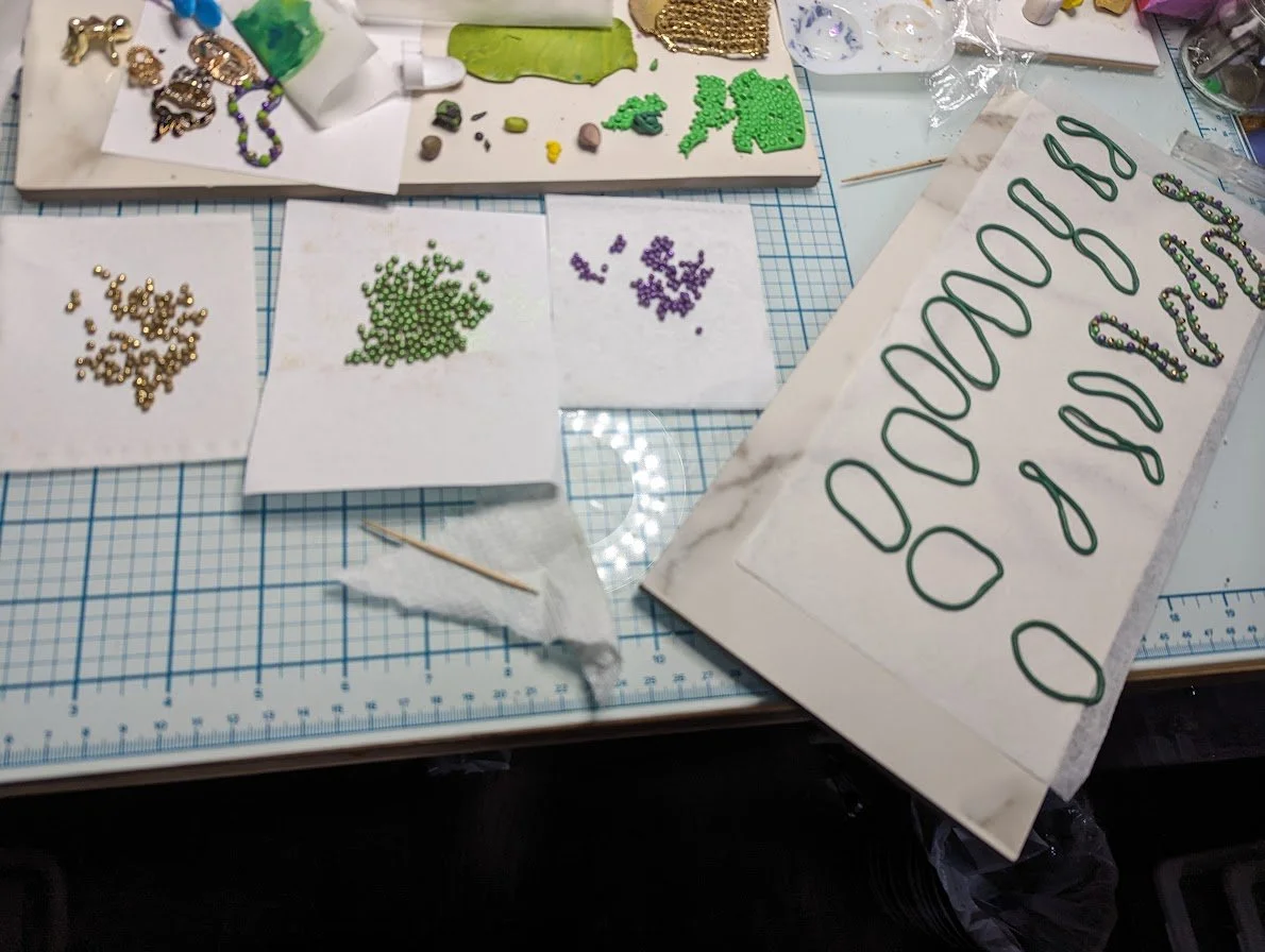 Studio workbench view of Mardi Gras King Cake Baby magnets/pins in progress by Art Illusions by Young. Hand-sculpted clay necklaces and colorful rolled clay beads and tools are arranged for scale, illustrating the detailed assembly process that creat