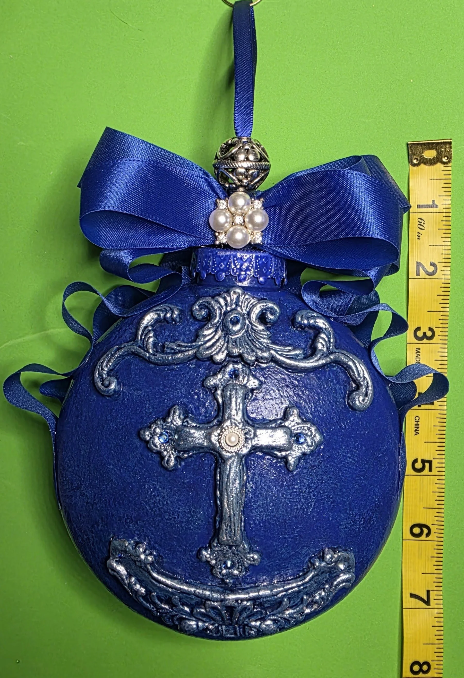 Front view of the Royal Blue Cross Ornament by Art Illusions by Young with ruler for scale. The luminous cobalt blue orb (approximately 7.5 inches in height) features an ornate silver cross with filigree and scrollwork, crowned by a sparkling royal-b