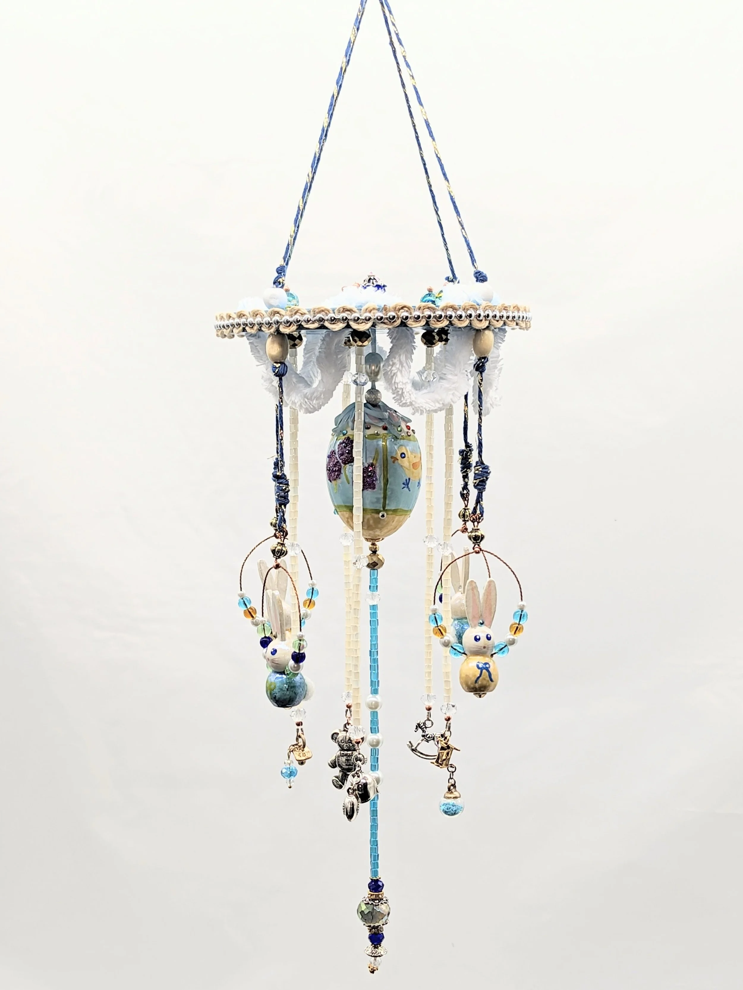Suspended view of the Baby Mobile – Cherished Nursery Keepsake by Art Illusions by Young. The six-inch blue wooden disc with scalloped white fluffy cording and golden accents anchors the composition, with hand-painted wooden bunnies, the decorated bl