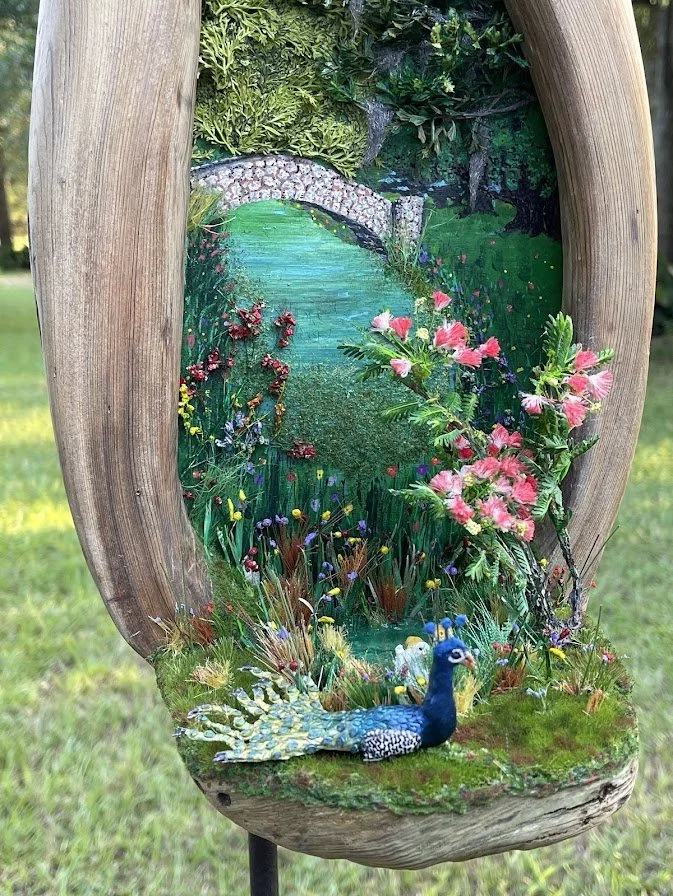 Close-up detail of the Enchanted City Park Serenity micro diorama showing the proud peacock, epoxy pond, miniature arched bridge, and vibrant pink mimosa flowers inside the hollow cypress tree trunk. Hand-sculpted and painted by Art Illusions by Youn