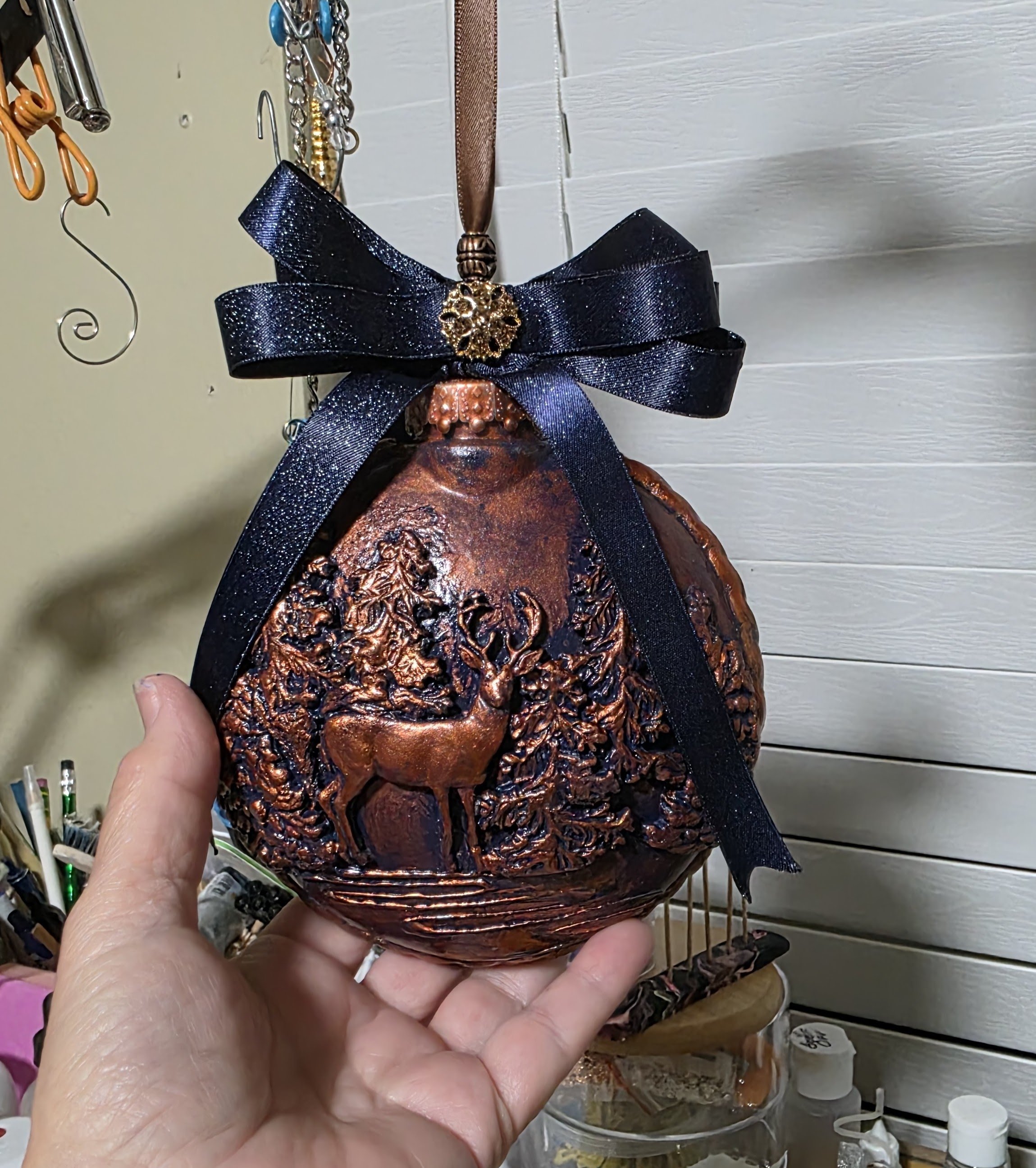 Close-up of the Woodland Majesty Ornament held by the artist, highlighting the hand-sculpted stag among textured pines and foliage. Rich copper-bronze metallic finishes bring warmth and depth to the relief work, while the navy-blue ribbon bow frames 