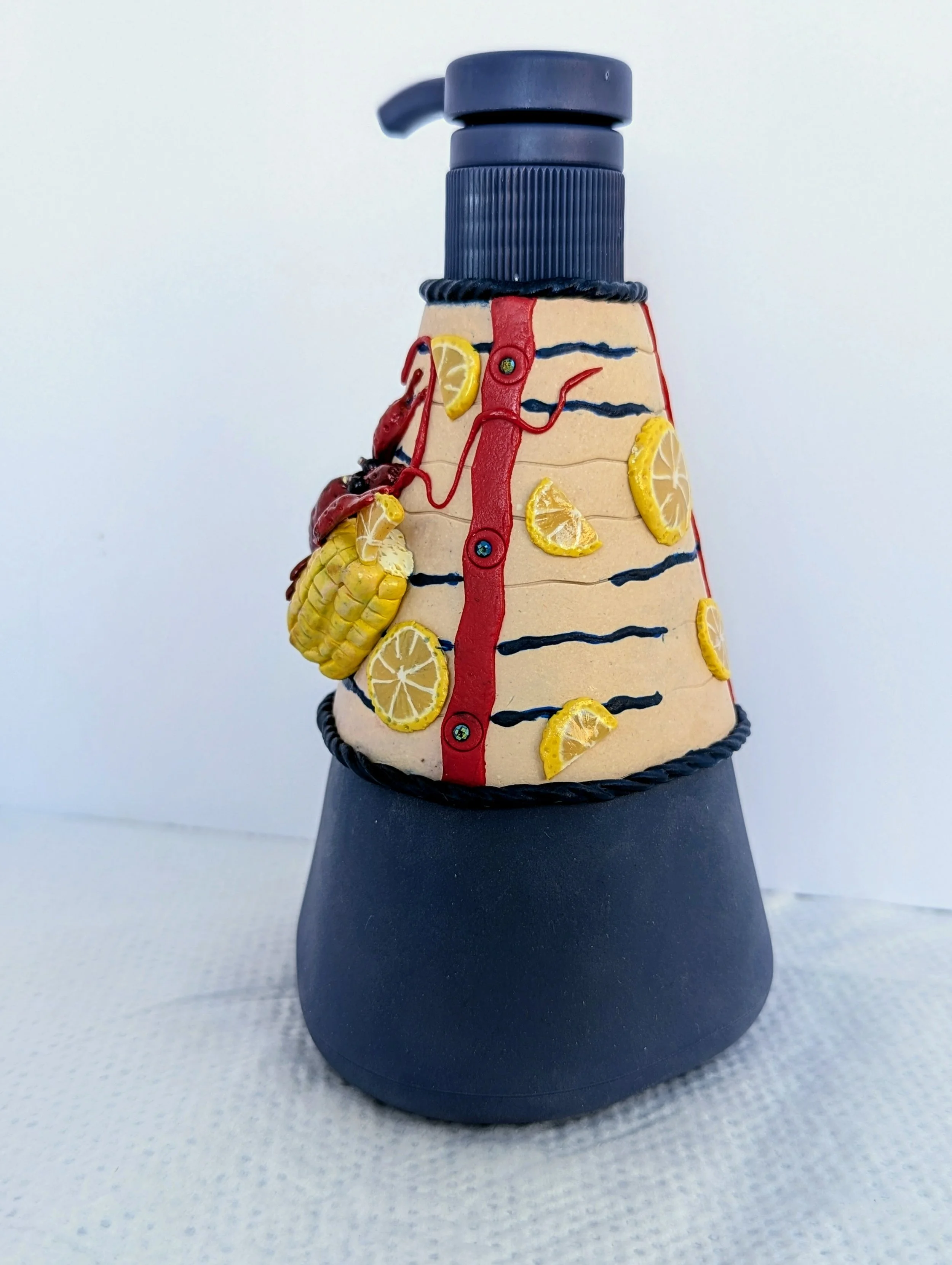 Angled view of the Crawfish Boil Soap Dispenser by Art Illusions by Young. The hand-sculpted polymer clay elements wrap gracefully around the triangular form, creating a three-dimensional scene of a classic Louisiana crawfish boil. Realistic red craw