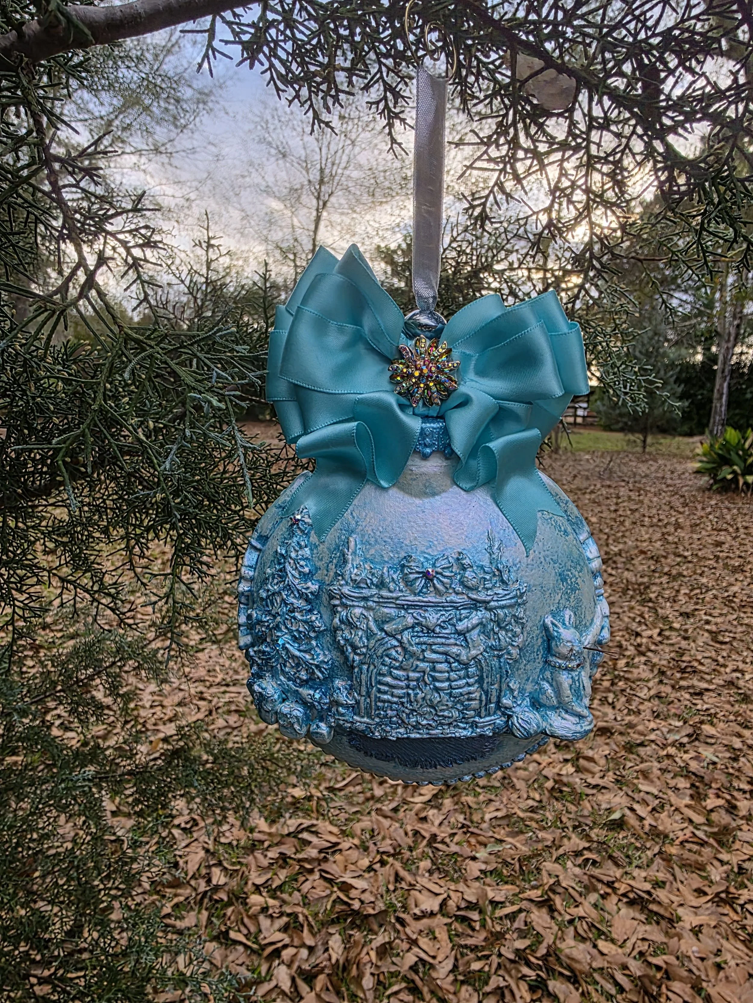Hanging frontal view of the Cozy Fireplace with Cat Ornament by Art Illusions by Young from Folsom, Louisiana. This hand-sculpted and molded spherical ornament features a rich, sparkling turquoise blue surface with delicate textured and subtle irides