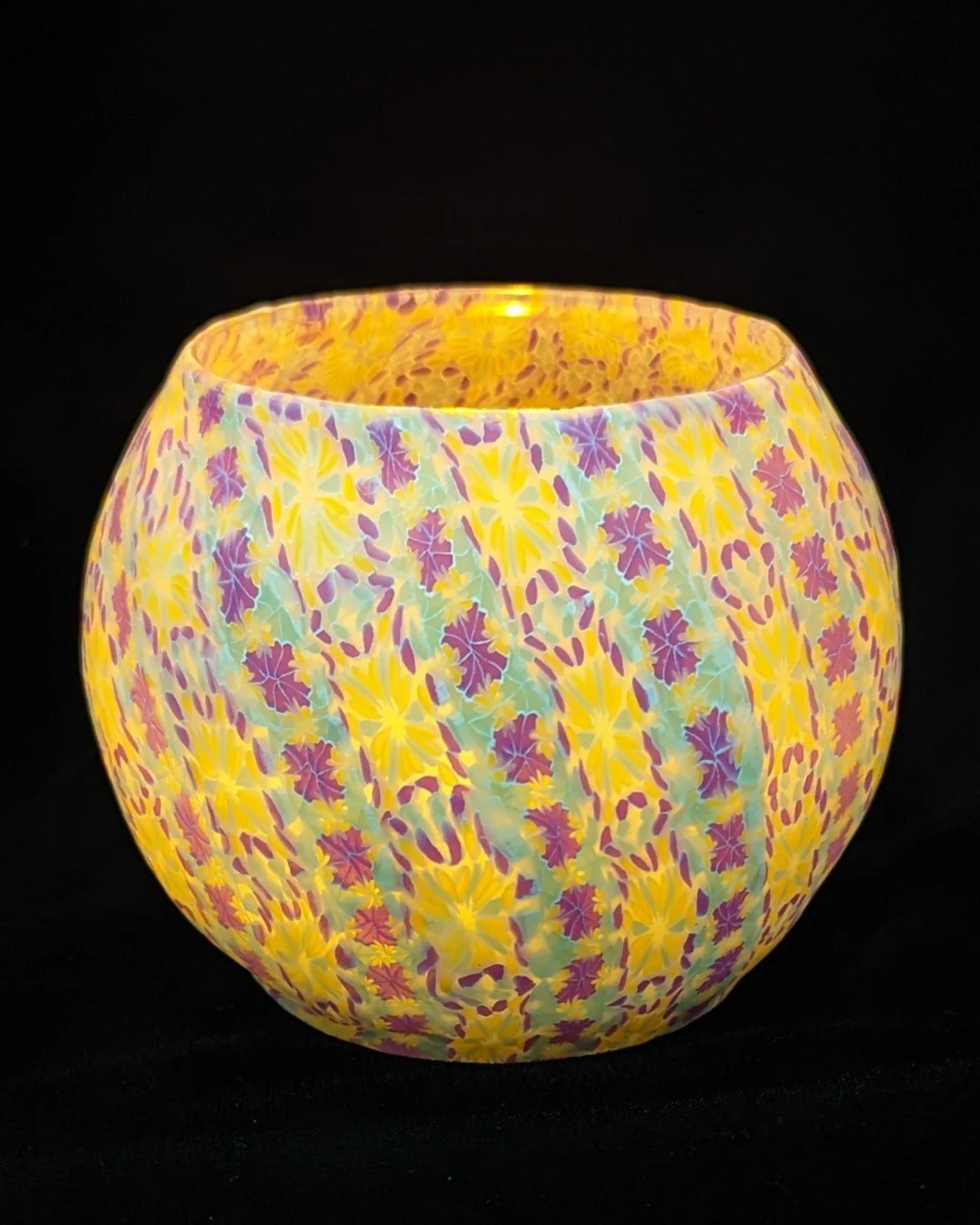 Translucent Floral Candle Holder illuminated against a dark background by Art Illusions by Young. When lit from within, the overlapping translucent polymer clay layers glow with warm, diffused light, revealing delicate floral patterns in soft purples