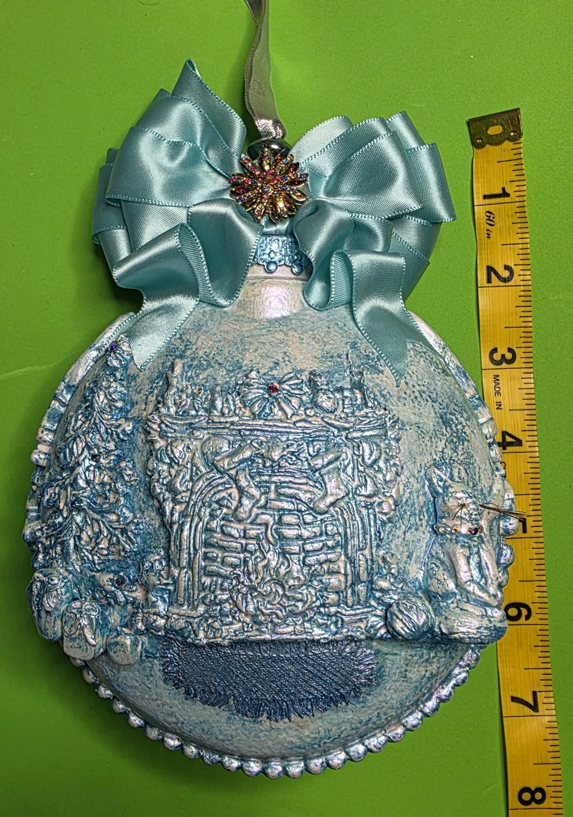 Close-up with vertical measurement of the Cozy Fireplace with Cat Ornament by Art Illusions by Young on a green background . The rich, sparkling texture in soft aqua-blue tones is highlighted, with the generous teal ribbon bow framing the composition