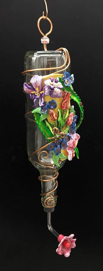 Hummingbird Feeder by Art Illusions by Young, Folsom, Louisiana. Recycled glass bottle adorned with hand-sculpted polymer clay irises and flowers in purple, pink, and blue tones, wrapped in decorative copper wire with a small red flower feeding tip.